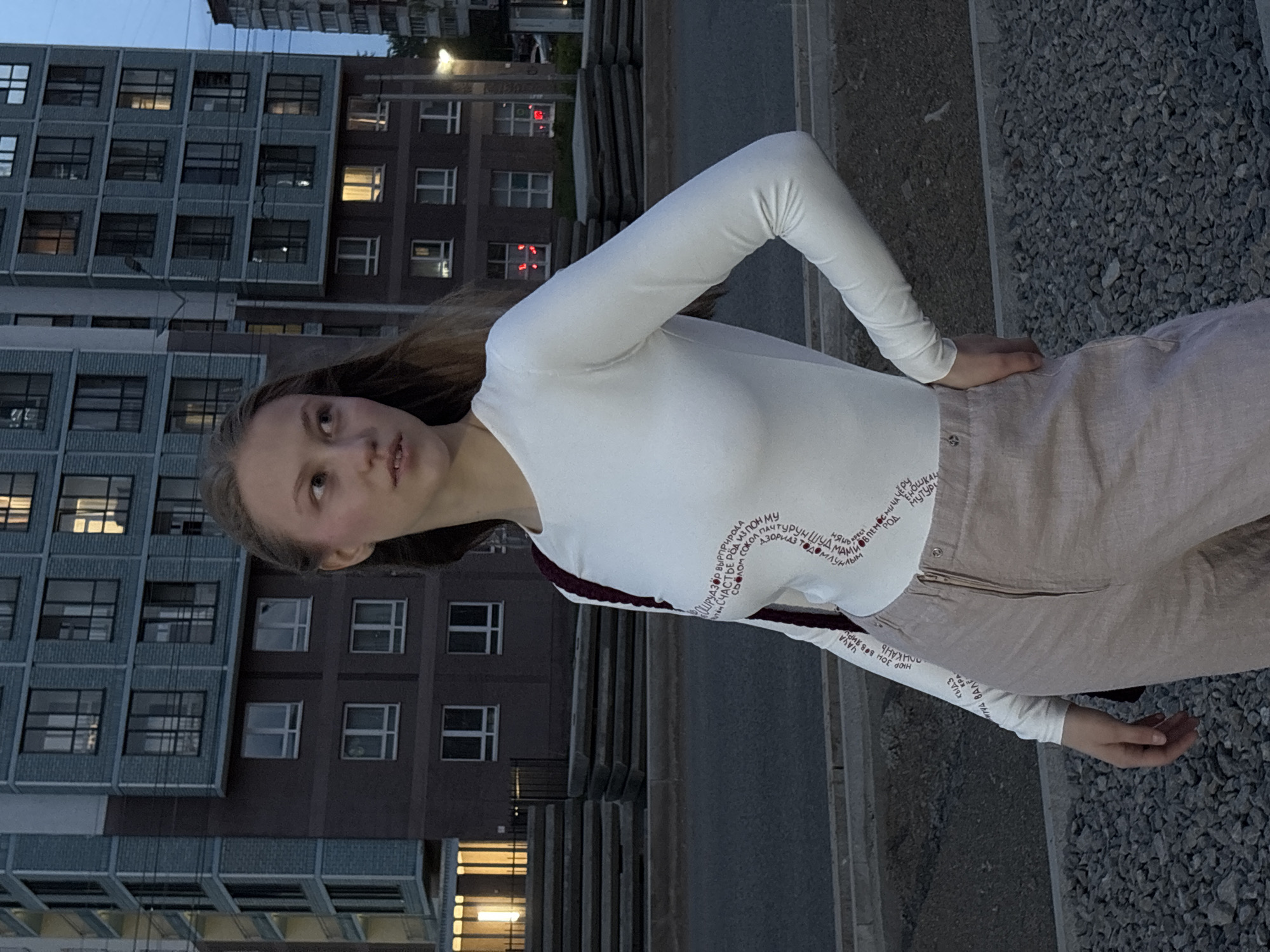 A fair-skinned, brown-haired woman stands angled toward the camera in what appears to be an urban setting. She is wearing a long-sleeved white top with red text bordering the front and back, and beige pants. The woman looks up and to the left of the frame.
Behind her, an apartment building with multiple windows rises vertically. The area around her appears to be paved, with a patch of gravel to her right. There are concrete steps behind her. The setting may be the city of Perm, Russia, based on the appearance of the architecture.