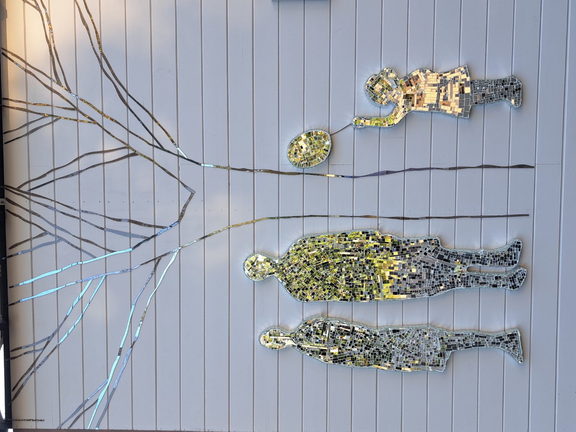 The image shows a mosaic artwork on a white, horizontally-paneled wall, likely in Irkutsk, Russia, although this cannot be confirmed from the image itself. The mosaic depicts a stylized tree with branches made of reflective, fragmented pieces of mirror. Beneath the tree, three human figures are represented in mosaic. On the left, two adult figures stand side-by-side. The figure on the left is taller and appears to be made entirely of mirror fragments. The figure on the right is shorter and wider, also composed of mirror fragments, with some parts reflecting a greenish hue, possibly from the surrounding environment. To the right of the tree trunk, a smaller, child-like figure is depicted. This figure is reaching upwards, holding onto a string attached to a balloon, which is also rendered in mosaic. The child's figure is made of mirror fragments, with some sections reflecting a greenish hue. The overall composition suggests a family group, possibly a father, mother, and child, under a tree, with the child releasing a balloon. The reflective mosaic pieces create a dynamic interplay of light and color, mirroring the environment around the artwork.