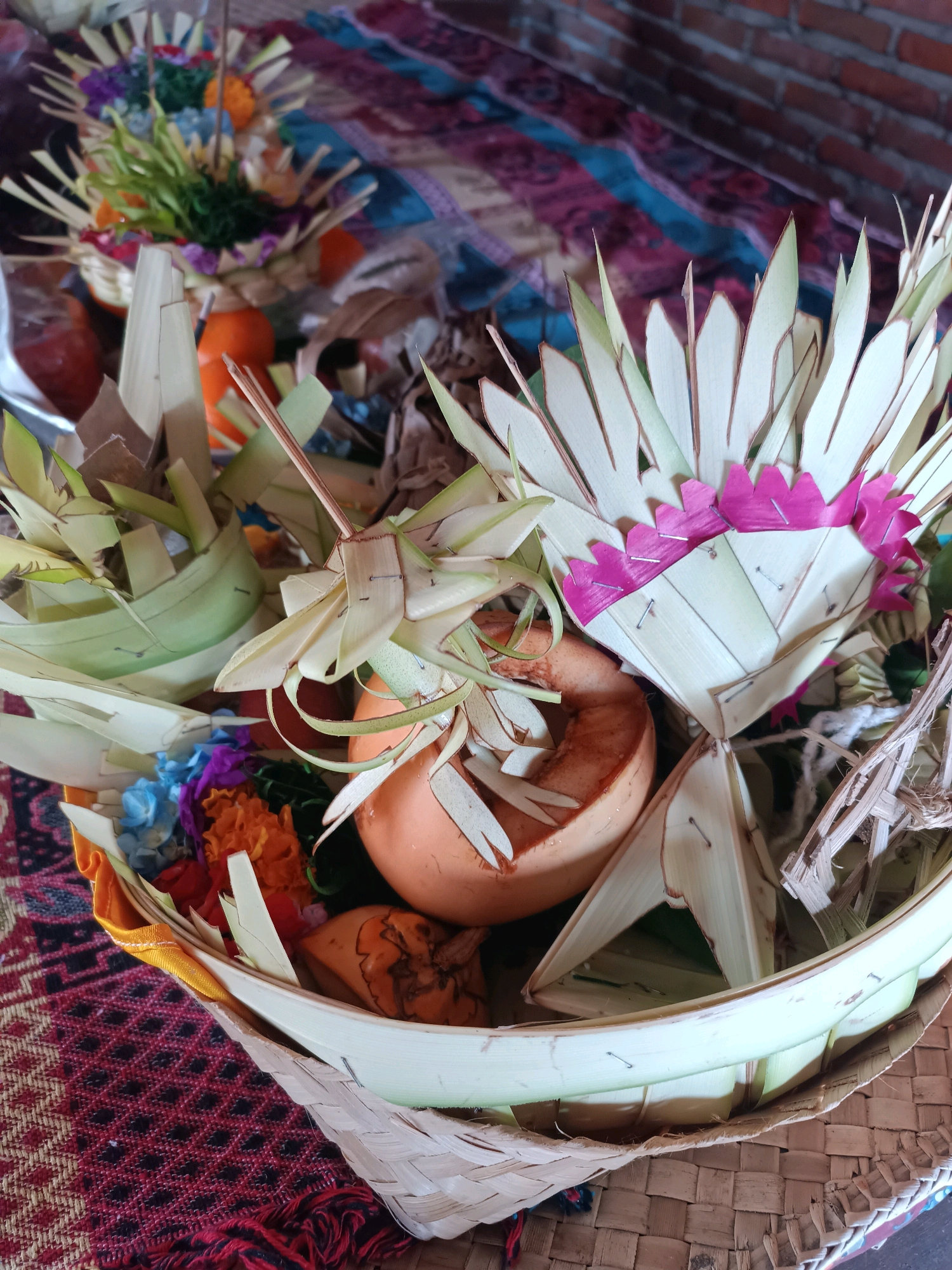 A close-up, eye-level shot captures an offering basket, likely a *canang sari*, in Selemadeg, Indonesia. The basket, woven from natural materials, is overflowing with various offerings. Prominently featured is a hollowed-out piece of orange fruit, possibly a pumpkin or gourd, placed centrally. Surrounding it are folded palm fronds arranged to create decorative elements, some edged with vibrant pink paper. Smaller, colorful artificial flowers in blue, purple, and orange are nestled within the arrangement, along with what appear to be dried leaves and twigs. The basket itself sits on a patterned textile, predominantly in shades of red and black, with a textured weave. In the background, out of focus, is a brick wall and another similar offering basket with a more distant view of fruit. The overall scene suggests a religious or cultural offering.