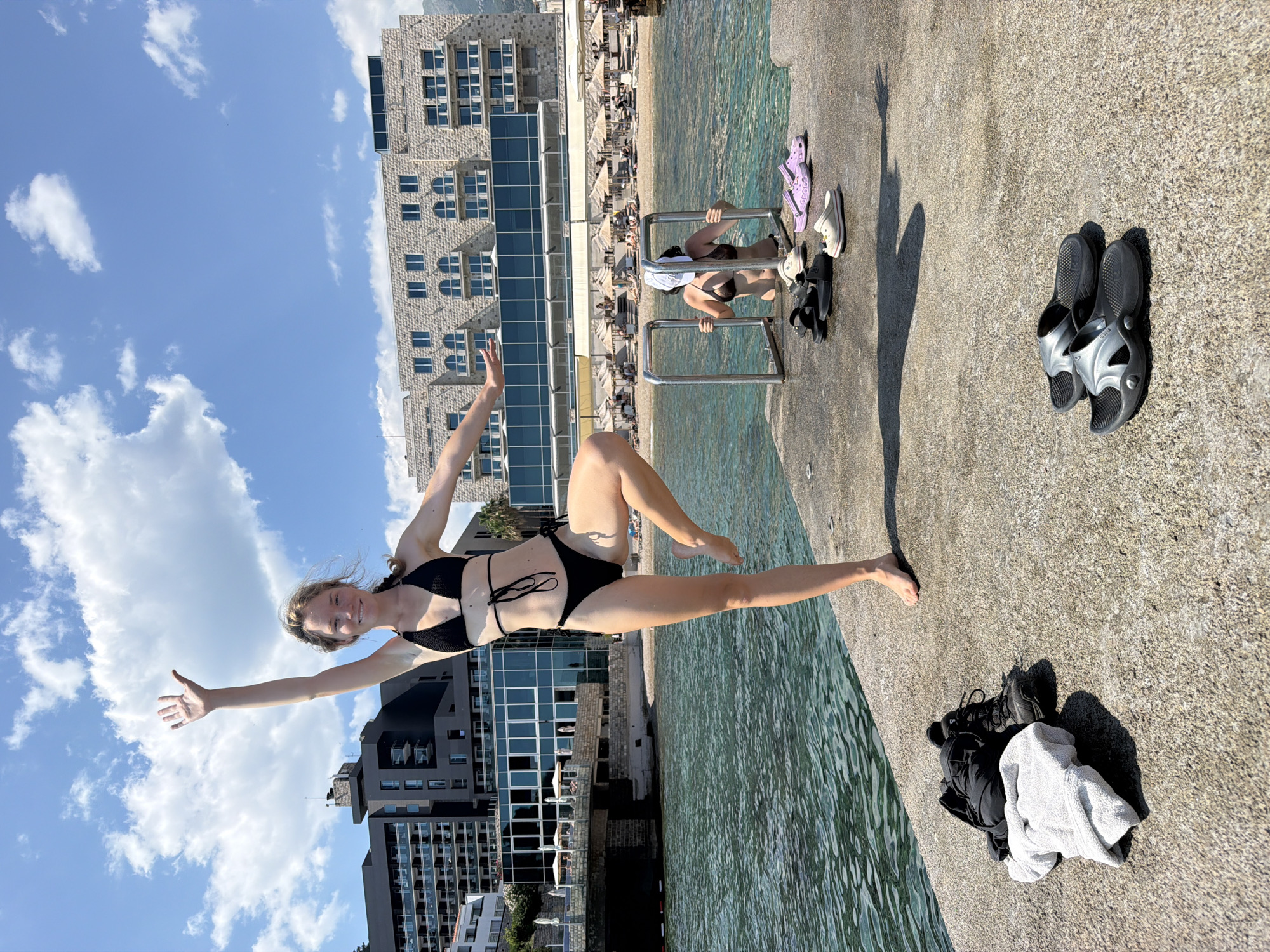 A young woman in a black bikini is captured mid-air, appearing to jump or leap near the edge of a pool or the sea. She is smiling and looking towards the camera with her arms outstretched, one hand giving a thumbs up. She is on a paved surface next to the water, which appears to be clear and blue.
In the background, there is a large, modern building with a stone facade and numerous windows. Beyond that, a beach with many loungers and umbrellas is visible, suggesting a resort or hotel setting. The sky is a clear blue with a few fluffy white clouds.
Several pairs of shoes and flip-flops are scattered on the pavement near the water's edge, including a pair of purple Crocs and some black sandals. A pile of dark clothing and a light-colored towel are also present. The sun is shining brightly, casting shadows on the ground, indicating it is daytime, likely late morning or early afternoon. The overall scene conveys a sense of fun, relaxation, and a vacation atmosphere, possibly in Budva, Montenegro, given the architectural style of the building and the coastal setting.