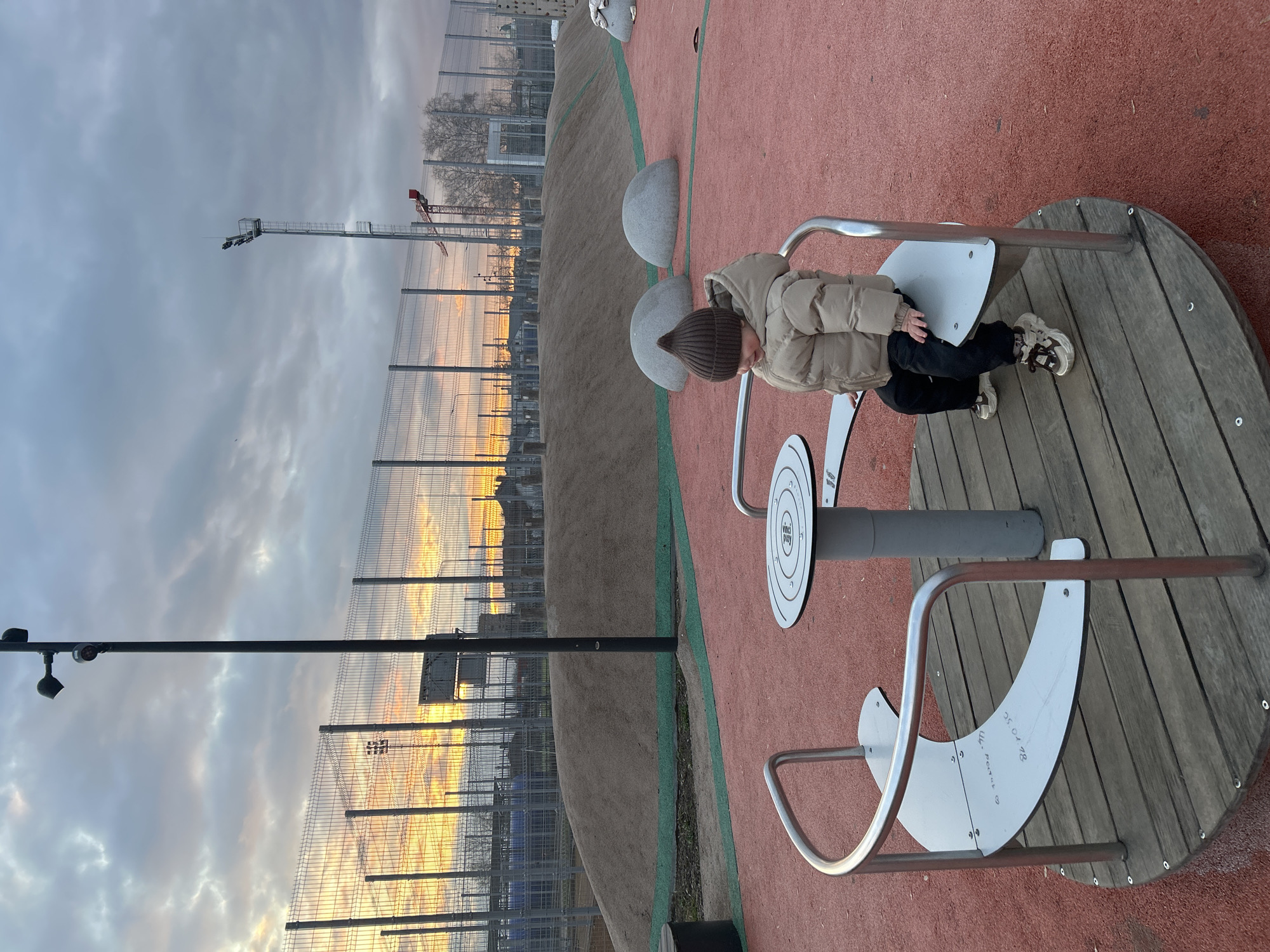 A child in a brown puffer jacket and beanie is sitting on a playground ride, with his head down. The ride is made of wood and has metal railings and seat-like attachments. The playground has a soft red surface. In the background, there is a tall fence, buildings, and a crane. The sky is cloudy with orange and yellow streaks near the horizon, suggesting sunset or sunrise. A street lamp is visible on the left. The ride appears to be a "Vinci Play" creation, according to text on its surface. The playground is in Klin, Russia.