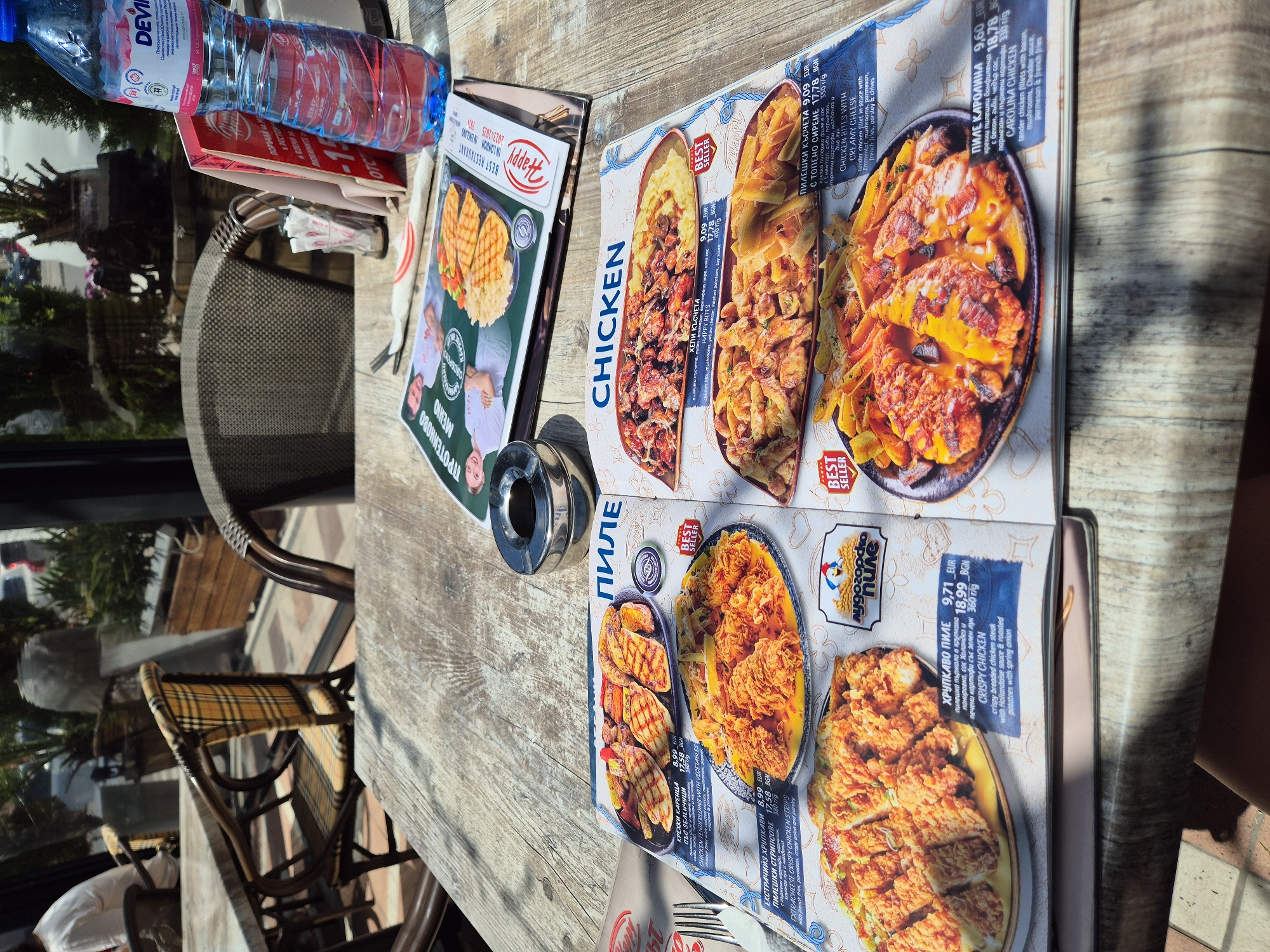 The image shows a table at an outdoor restaurant. On the table are a menu opened to the chicken section, a water bottle, a restaurant bill and receipts, an ashtray, and cutlery. The chairs around the table are made of brown metal and have woven seats. The floor is tiled in a brick pattern. Other tables and chairs can be seen in the background, as well as trees and buildings.