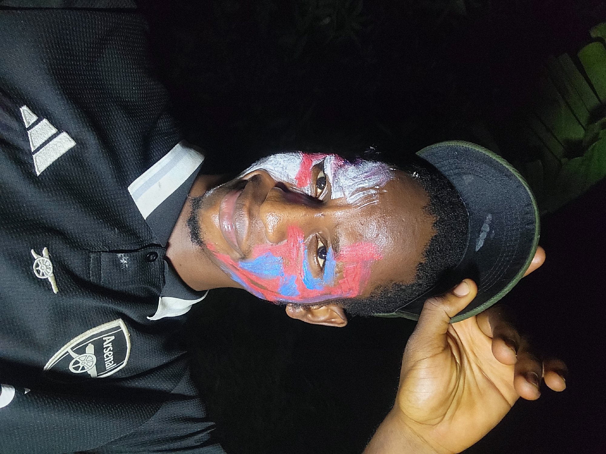 A man with dark skin and short, dark hair wearing a black cap and a black Arsenal soccer jersey with white trim is photographed at night in a low-light environment. His face is decorated with white, red, and blue paint. The location is Nkpor, Nigeria.