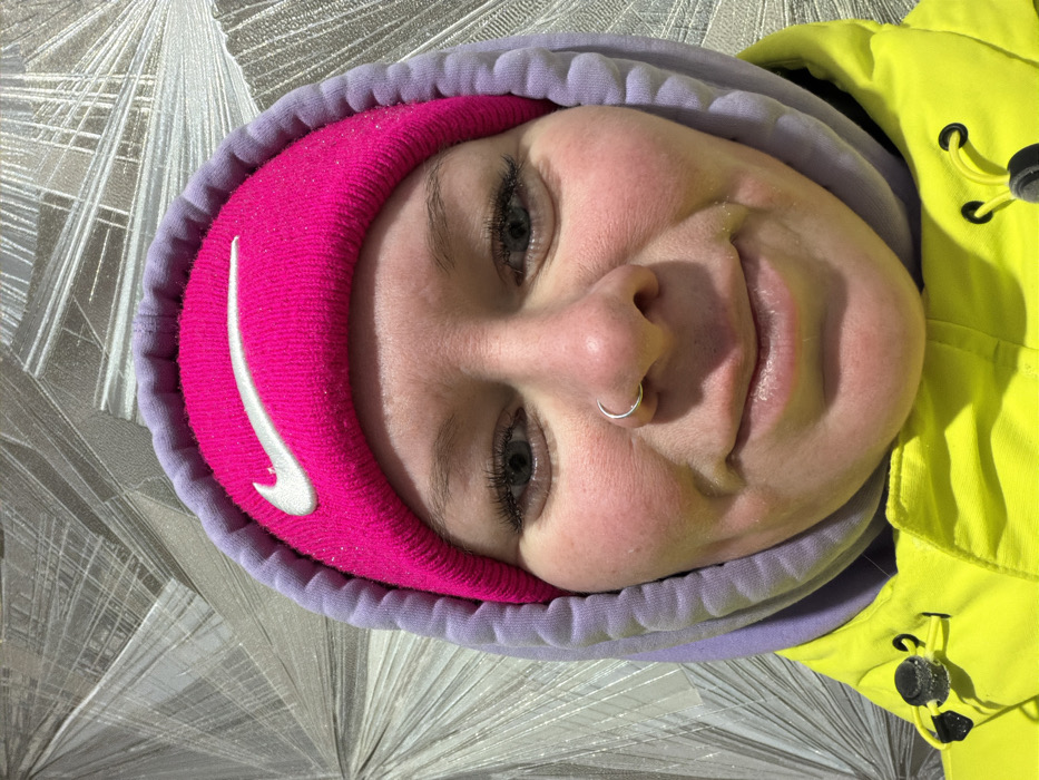 The image displays a close-up portrait of an individual, positioned centrally. The subject is wearing a bright pink knitted beanie hat, featuring a white Nike swoosh logo on the front, which is partially covered by the hood of a light purple sweatshirt. Small, reflective particles are visible on the beanie. Below the sweatshirt, a bright yellow outer jacket is visible, with black and yellow toggles on a drawstring near the collar. The individual has fair skin with flushed cheeks, blue-grey eyes with visible eyelashes, and a silver hoop nose ring in the left nostril. A slight smile is present, and the subject is looking directly towards the viewer. The background consists of a silvery-grey wall with a repeating geometric pattern of radiating lines. The scene is located in the city of Chaikovsky, Russia.