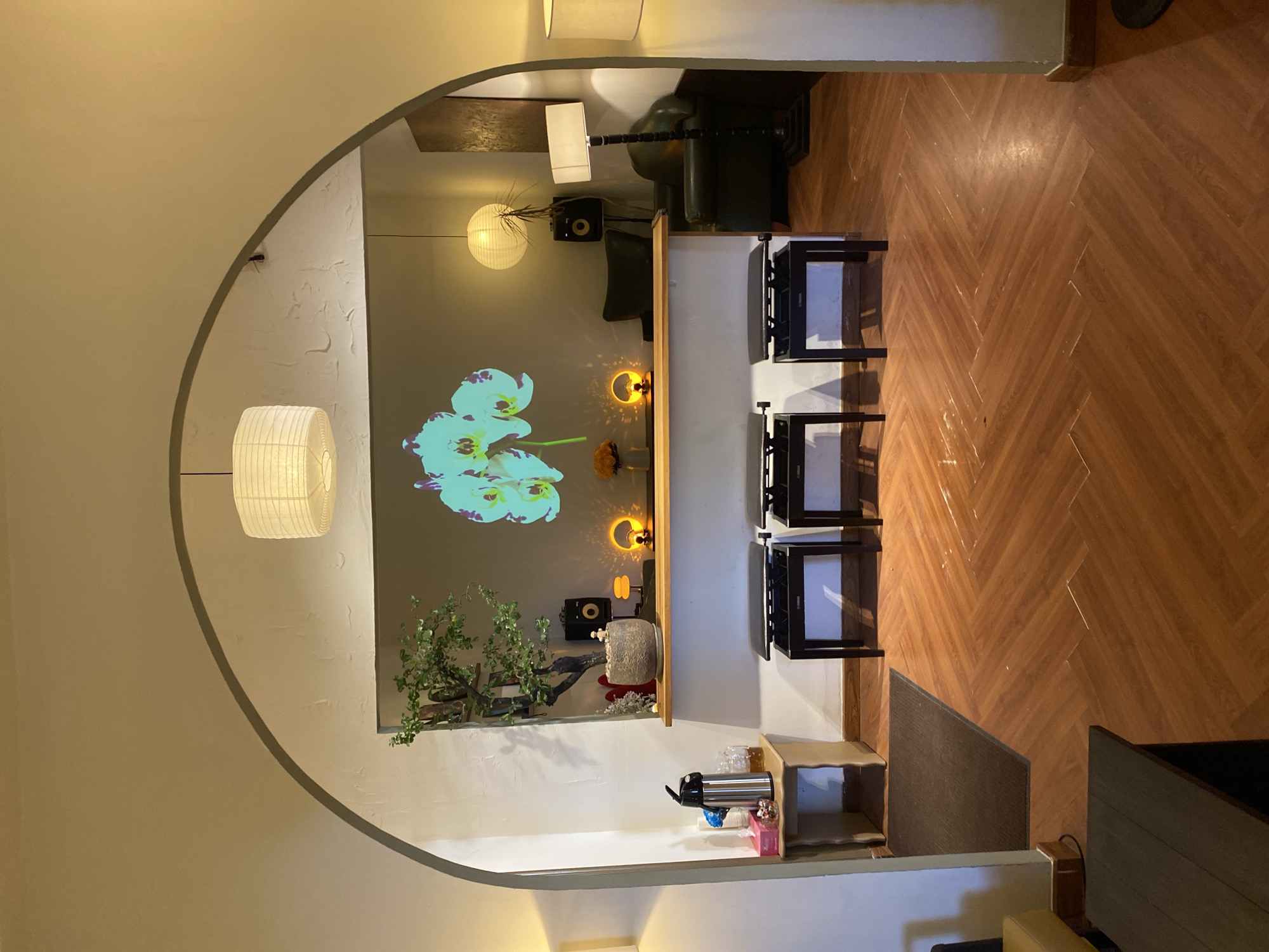 The image depicts an indoor scene, likely a lounge or waiting area, characterized by warm lighting and an arched entryway. In the foreground, a wooden floor with a herringbone pattern is visible. Three black piano benches are arranged side-by-side in front of a white counter.
Above the counter, a projector displays a large image of a light blue orchid against a grey wall. To the left of the projected image, a potted bonsai tree sits on a shelf, accompanied by a small figurine. Behind the bonsai, a speaker is mounted on the wall. Two small, round lamps with orange light are placed on the counter, casting a warm glow.
Further back, to the right of the projected orchid, a green armchair is visible, and next to it stands a tall, dark floor lamp with a white shade. Another round, white lantern hangs from the ceiling near the center of the room. On the far right, partially obscured by the archway, a portion of another white lamp is visible.
To the left of the counter, a small side table holds a stainless steel thermal carafe, a pack of tissues, and some small bowls containing what appear to be candies or snacks. A dark brown mat lies on the floor in front of this table. The overall ambiance suggests a relaxed and inviting atmosphere, possibly within a cafe, a music studio, or a similar establishment in Lâm Viên - Đà Lạt, Vietnam. There are no people visible in the image.