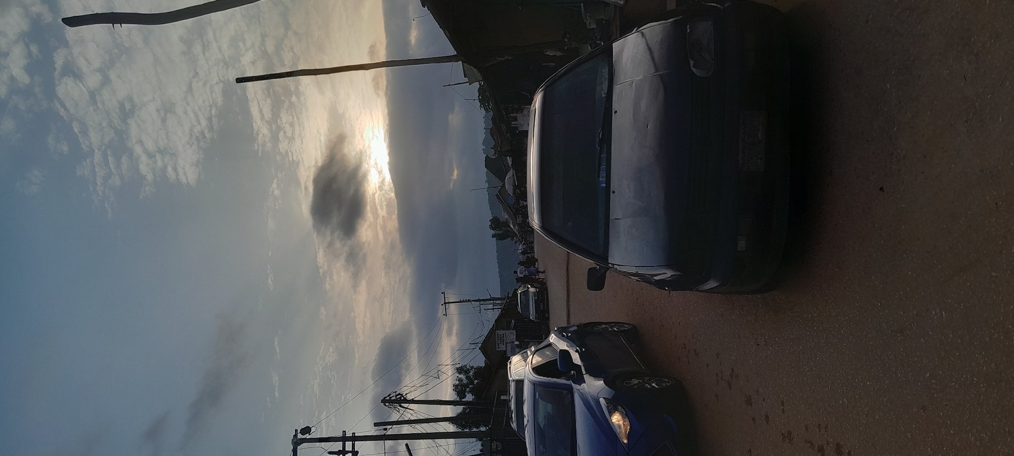 The image depicts a street scene in a town or city, possibly Abuja, Nigeria, at dusk. The sky is overcast with dramatic clouds, and the sun is setting, casting a warm glow through the clouds. Several vehicles are present, including a dark-colored car in the foreground, a blue hatchback on the left, and other cars further down the street. Buildings line the sides of the road, with some appearing to be shops or homes. Utility poles and power lines are visible, suggesting an urban or semi-urban environment. A few people are seen walking and interacting along the street, and there is a sense of activity. The overall atmosphere is one of a community winding down for the day.
