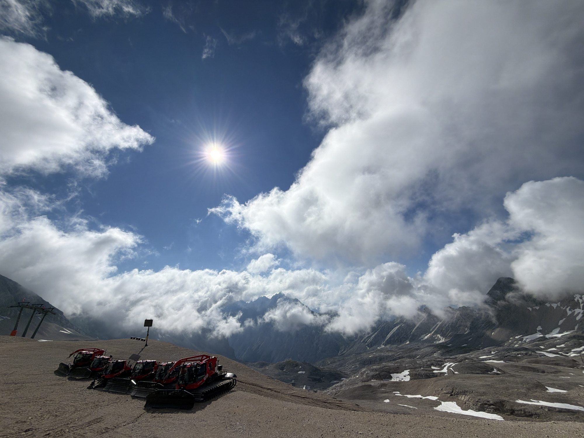 This image depicts a mountainous, rocky landscape under a bright, sunny sky with scattered white clouds. In the foreground, a line of five red snowcats is parked on a gravelly surface. To the left, a ski lift structure with three towers is visible. In the background, rugged, snow-dusted mountains rise, partially obscured by clouds. A dark signpost stands behind the snowcats. The scene appears to be located in a high-altitude area, likely a ski resort or a mountain pass, possibly in the vicinity of Garmisch-Partenkirchen, Germany, given the presence of ski infrastructure and the alpine environment.