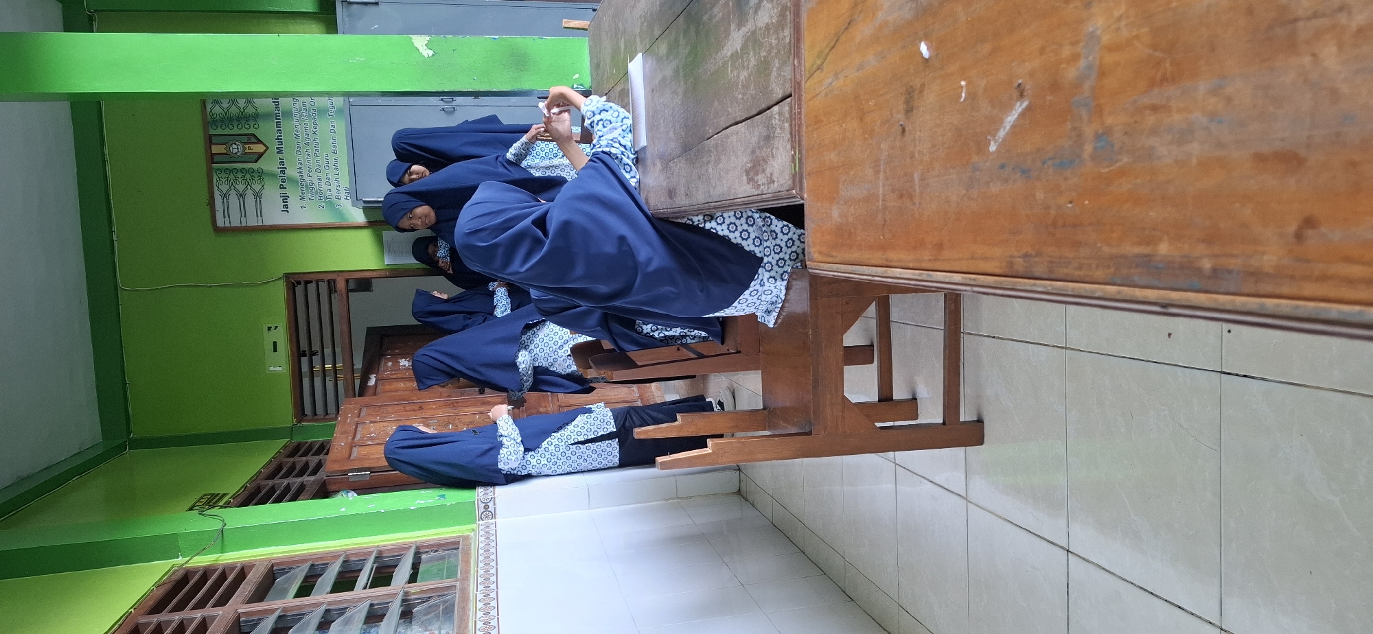 The image depicts a classroom in the city of Wonorejo, Indonesia. Several young female students, dressed in navy blue hijabs and patterned blouses, are gathered around a wooden table. Some are standing, while others are seated on wooden chairs. One student in the foreground appears to be holding up a phone, possibly taking a photo or video. The room is brightly lit with green walls and white tiled floors. A wooden door and a window with louvers are visible on the left side of the frame. A poster with text and a logo is displayed on the green wall. The atmosphere suggests a casual gathering or break time for the students.