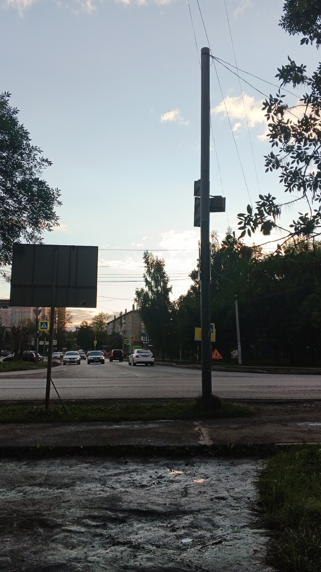 The image shows a street scene in Izhevsk, Russia, likely during the evening or early morning due to the low light and soft sky. In the foreground, a wet, muddy patch of ground with tire tracks and reflections is visible, suggesting recent rain or a drainage issue. A dark-colored sidewalk runs along the edge of this patch.
Dominating the right side of the frame is a tall, gray utility pole with electrical wires extending across the sky. A traffic signal or light fixture is attached to the pole. On the left side of the image, a large, dark rectangular billboard or sign is partially visible, mounted on a pole.
The middle ground depicts a multi-lane road with several cars driving in both directions. The cars appear to be a mix of sedans and SUVs. Pedestrian crossing signs are visible on the roadside. Trees and buildings line the street, with apartment buildings in the distance and dense foliage closer to the road. The sky above is a pale blue with scattered white clouds, and the sun appears to be low on the horizon, casting a warm glow.
The overall impression is a typical urban street in Izhevsk, Russia, captured during a period of low natural light.