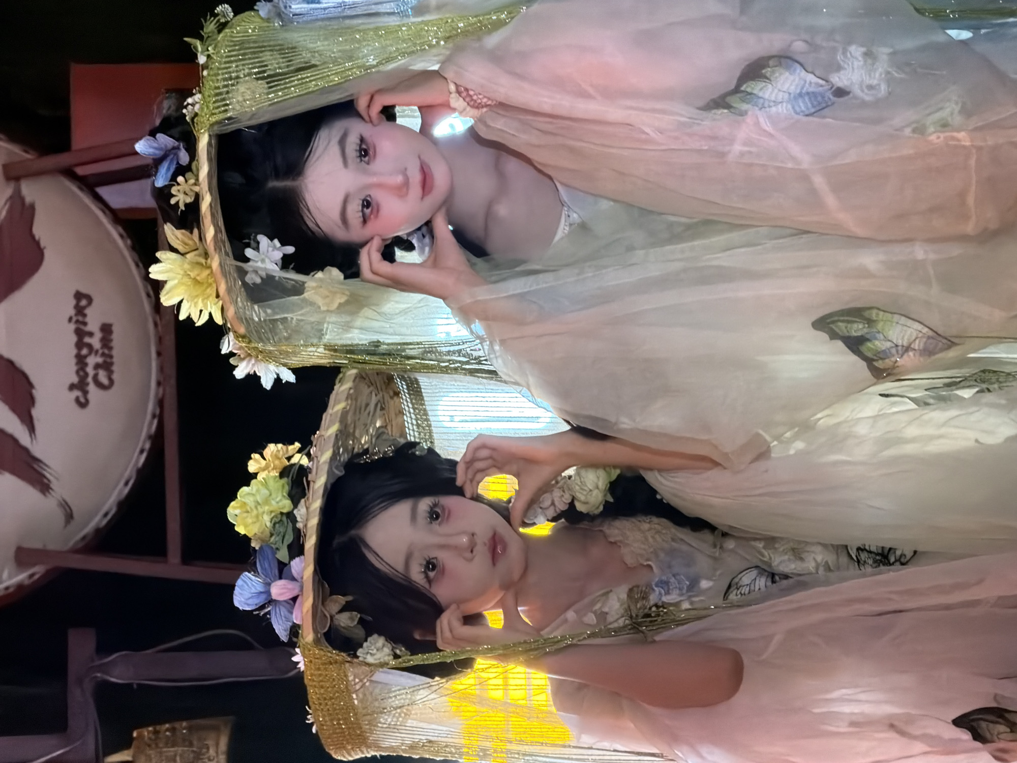 The image shows two young East Asian females dressed in traditional Chinese attire with elaborate headwear adorned with flowers and butterflies. They are positioned in the foreground, with their faces close to the camera, each touching their cheeks with their hands. The female on the left wears a light-colored, possibly silk, garment with floral embroidery and a straw hat-like headdress decorated with gold trim and greenery. The female on the right is similarly adorned, with a sheer, light pink garment over an inner layer, and a headdress featuring light green netting and flowers. In the background, a large round object, possibly a drum or a decorative element, is visible with Chinese characters that read "Chongqing China." The overall setting appears to be a decorative, possibly indoor, display or performance space, consistent with cultural exhibitions or events in Yuzhong District, Chongqing, China. The lighting is dramatic, with soft, warm tones illuminating the subjects.