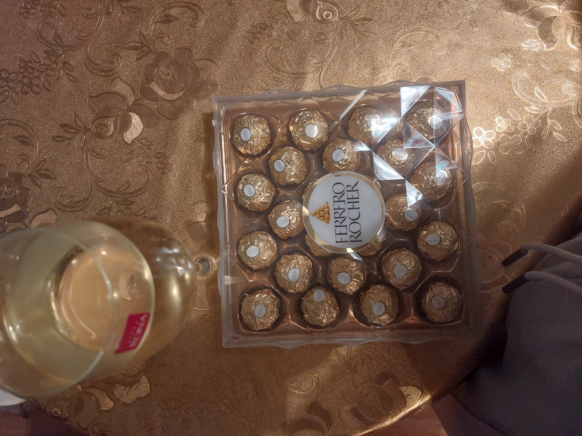 A box of Ferrero Rocher chocolates sits on a gold patterned surface, likely a tablecloth. Next to the box is a glass candle holder with a red label. A portion of grey clothing is visible on the right edge of the image. The location is unknown, the city of Tsarevo, Bulgaria is not visible.