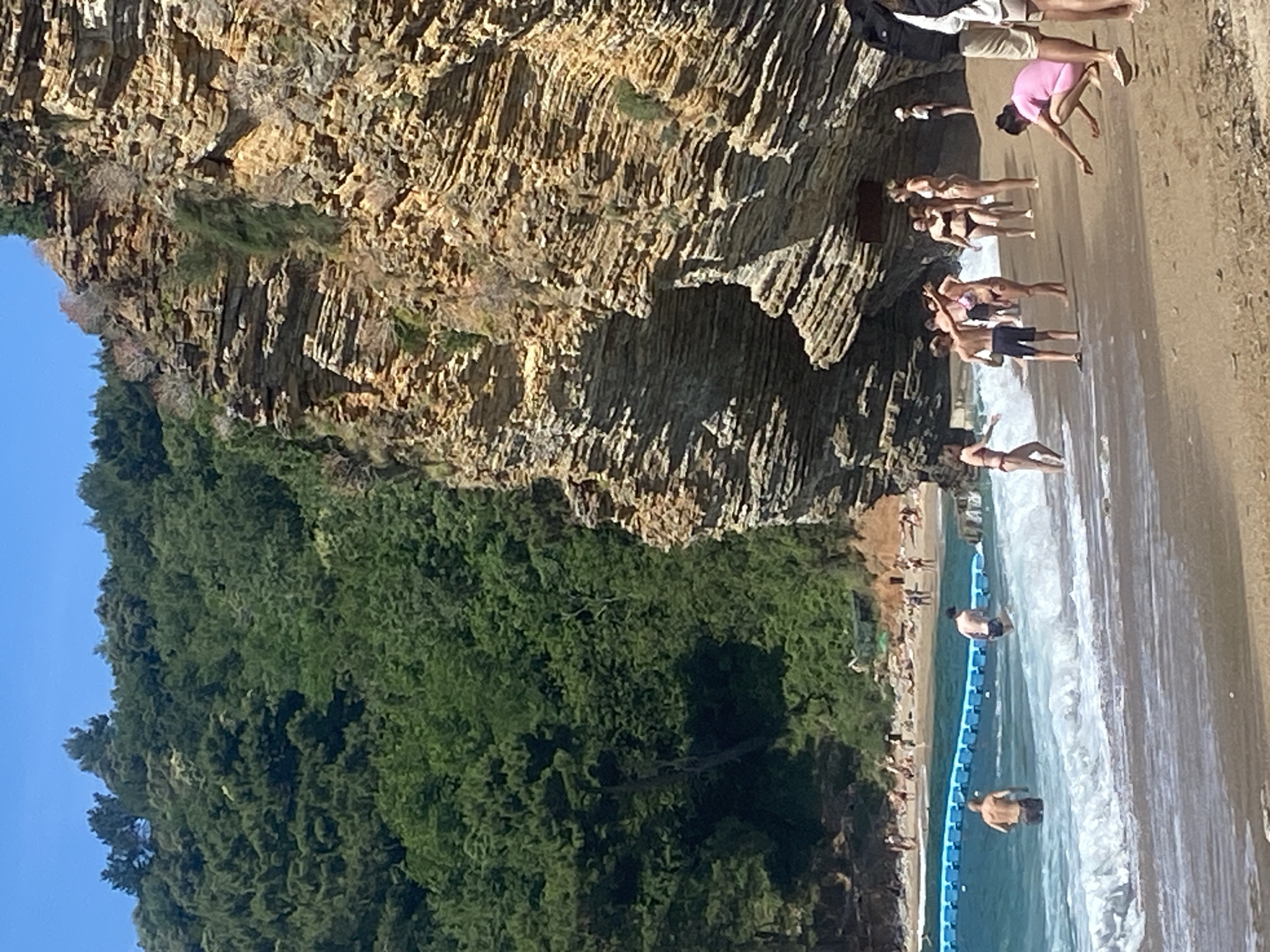 The image depicts a vibrant coastal scene at Sveti Stefan, Montenegro, with a dramatic cliff face, a sandy beach, and clear blue water. The photograph is oriented vertically, showing the cliff on the left, the beach and sea on the right, and a clear blue sky at the top left corner.
The most prominent feature is a large, rugged rock cliff, characterized by horizontal layering and varying shades of brown and grey, indicating geological strata. Lush green trees and vegetation cover the upper part of the cliff and extend beyond it, contrasting with the bare rock.
At the base of the cliff, a sandy beach stretches along the coastline. Numerous people are present on the beach and in the water, enjoying what appears to be a sunny day. Many individuals are dressed in swimwear, with some standing on the wet sand, others wading in the shallow water, and a few seemingly stretching or relaxing. One person in a pink top and light shorts is bending forward on the sand directly under the cliff. Another group of people, also in swimwear, stands further back on the beach, near the cliff's edge.
A blue floating barrier, composed of interconnected buoyant elements, is visible in the water, demarcating a section of the swimming area. Waves gently break on the shore, creating white foam. The water appears clean and inviting, reflecting the bright sunlight. The overall impression is one of leisure and natural beauty. There is no visible text.