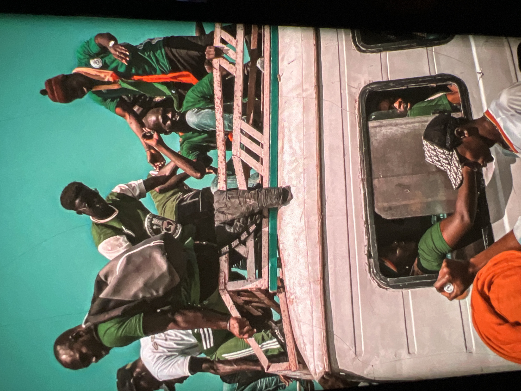 The image captures a vibrant scene with multiple individuals packed onto the top and inside a vehicle, likely a bus or truck. The people on top are predominantly young men wearing green shirts, some with backpacks, suggesting a shared activity or event. One man in the foreground on top is wearing a brown beanie and a green shirt with a red and orange scarf, appearing to be gesturing or interacting with someone. Another man is seen with a grey backpack over his green shirt. The vehicle itself is a light-colored van or bus, with a weathered exterior.
Inside the vehicle, through a window, a woman in a green shirt is visible. In the foreground, below the window, two young men are present. One is wearing a white shirt with blue and red trim and a black baseball cap with a white pattern. The other person's face is partially obscured, but they are wearing an orange head covering and a white shirt. The background is a bright, solid turquoise or teal color, which could represent a backdrop or possibly the sky.
While the image is rich in detail, there is no specific visual information within the frame that definitively places the scene in Lagos, Nigeria. The style of clothing, the vehicle, and the general atmosphere are common in many urban environments in Africa.
