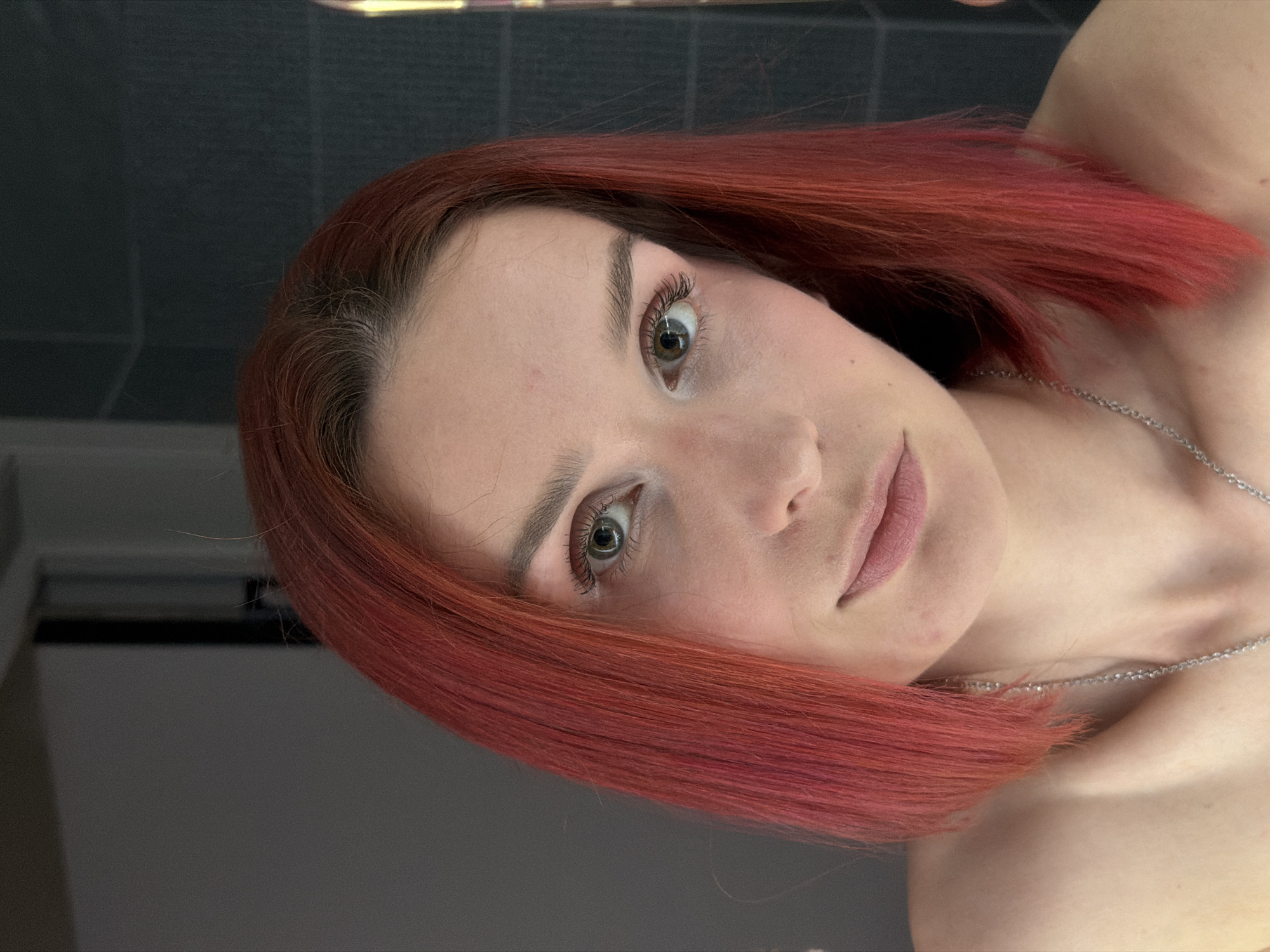 A close-up, slightly rotated, eye-level shot shows a fair-skinned woman with short, reddish-brown hair. She has green eyes, well-defined eyebrows, and light pink lips. A delicate silver necklace is visible around her neck. The background includes a dark tiled wall and part of a light-colored doorframe. The image provides no indication of location, thus nothing indicates Chaikovsky, Russia.