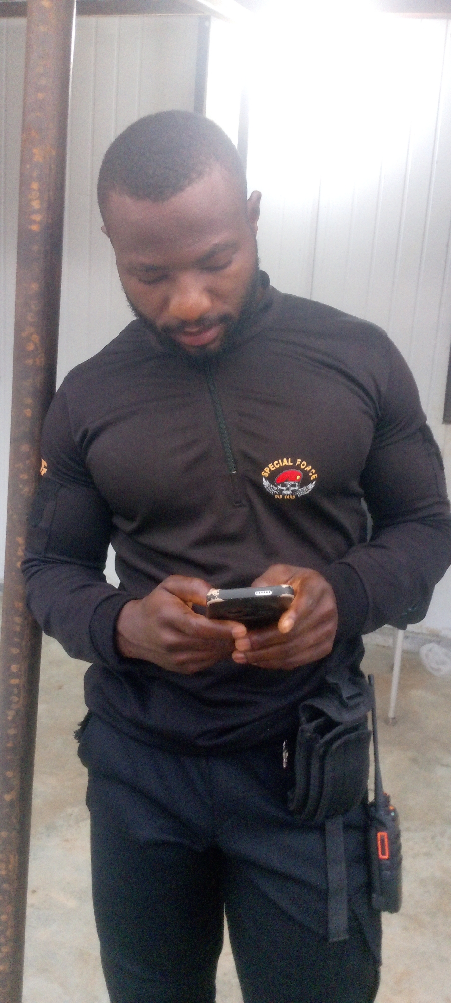 In Indo, Nigeria, a Black man with a shaved head and beard is standing next to a rusty pole. He is wearing a black long-sleeved shirt with a zipper at the neck and a patch on the left chest that reads "SPECIAL FORCE DIE HARD". He also has black pants and a tactical belt with a radio attached to the right side. The man is looking down at a smartphone in his hands. The background appears to be an indoor area with a white, corrugated wall and bright lighting.