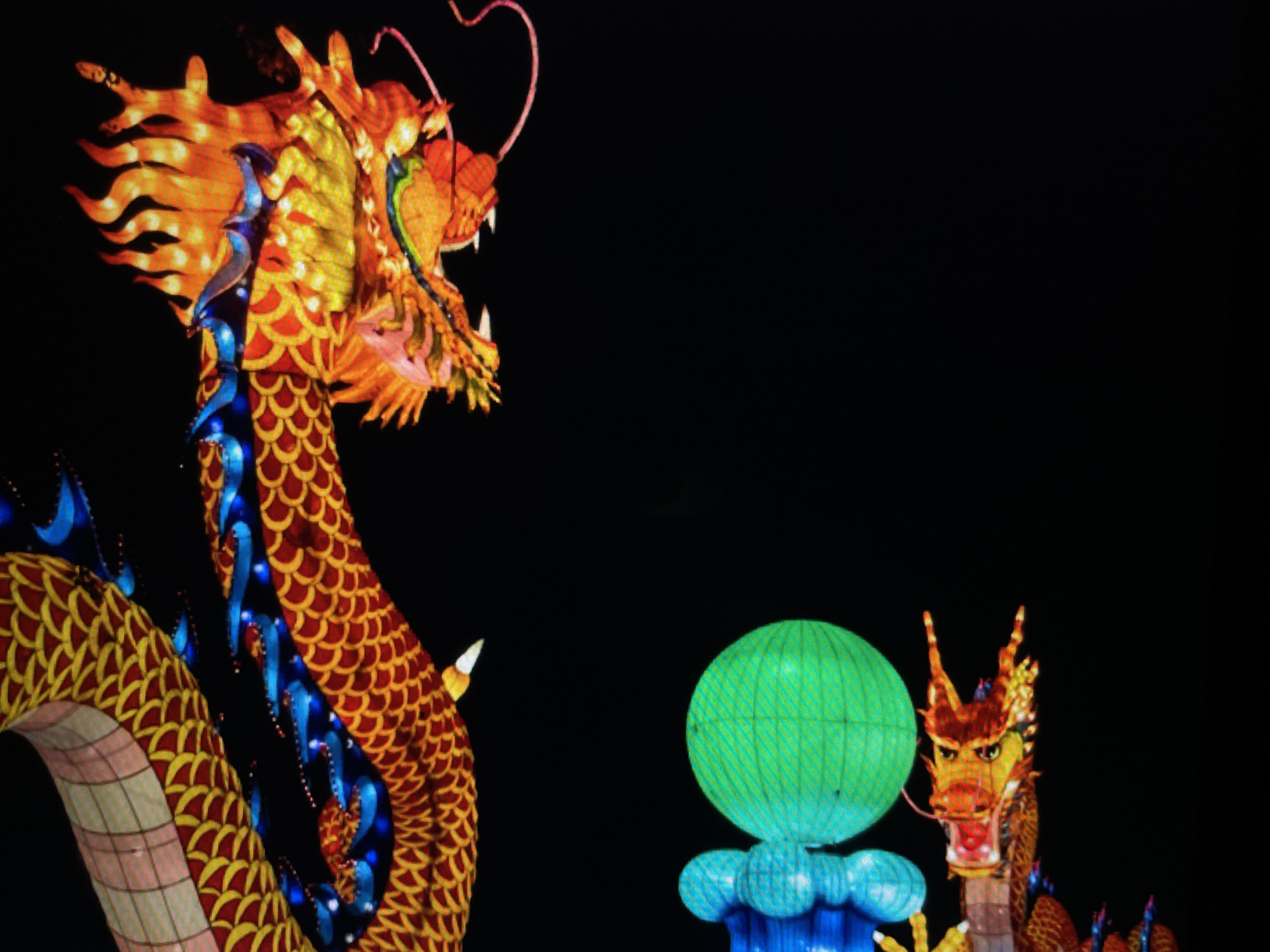 Two Chinese dragons are illuminated against a black background. The dragon on the left is large and detailed with a yellow and red scaled body and an orange head. It has blue highlights and a fierce expression. The dragon on the right is smaller and stands near a green globe with a blue base. It has a red and yellow head with horns. The scene is set at night. The location in the city is unknown.
