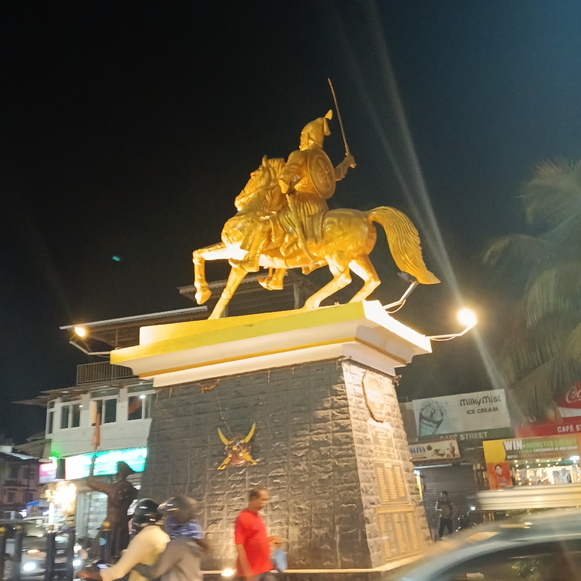 Captured at night, the image features a golden statue of a man riding a horse, holding a sword and shield. It stands on a stone pedestal with decorative elements, including a helmet. The statue is brightly lit, contrasting with the dark sky. The location appears to be a commercial area in Oxel, India, evident from the surrounding buildings with signages, including "Milky Mist Ice Cream" and "Cafe Street." There are people walking on the sidewalks, and vehicles are visible on the road. The scene suggests a bustling urban environment.