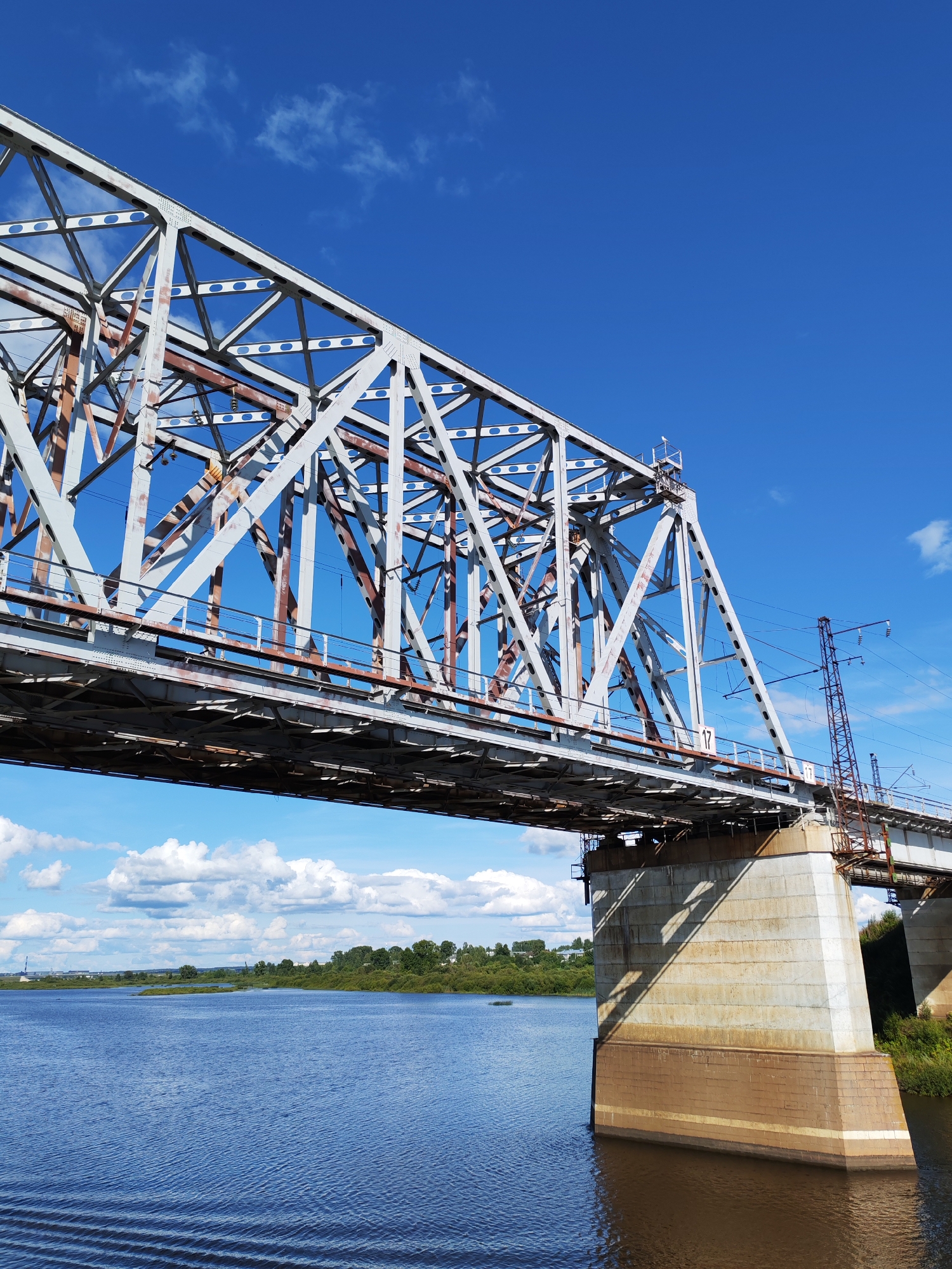 The image shows a railway bridge spanning across a wide river. The bridge is constructed of metal trusses and appears to be aged, with visible signs of rust. The river is blue and calm, reflecting the bright blue sky and scattered white clouds. The banks of the river are lined with green trees and vegetation. In the distance, there are buildings visible, suggesting a populated area. A support pillar of the bridge, made of concrete and stone, is prominently visible on the right side of the image, with shadows cast by the bridge structure. Utility poles and wires are also visible along the bridge. The scene is a daytime view under clear weather conditions. The location is identified as Tarkanovo, Russia.