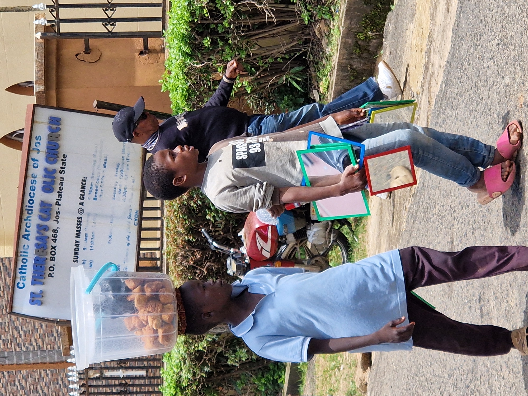 Two young boys and an adult male are walking on a paved street. The boy on the left is carrying a large clear plastic bucket filled with fried food items on his head. He is wearing a light blue collared shirt and dark brown pants. The boy in the center is wearing a gray t-shirt with "SPACE 93" printed on it and blue jeans. He is holding several small framed mirrors of various colors. The adult male is wearing a black baseball cap, sunglasses, a black sweatshirt, and blue jeans. He is walking behind the boys. In the background, there is a red motorcycle, lush green bushes, a black metal fence, and a building with a sign that reads "Catholic Archdiocese of Jos ST. THERESA'S CATHOLIC CHURCH P.O. BOX 468, Jos - Plateau State." The environment appears to be an outdoor, urban setting.