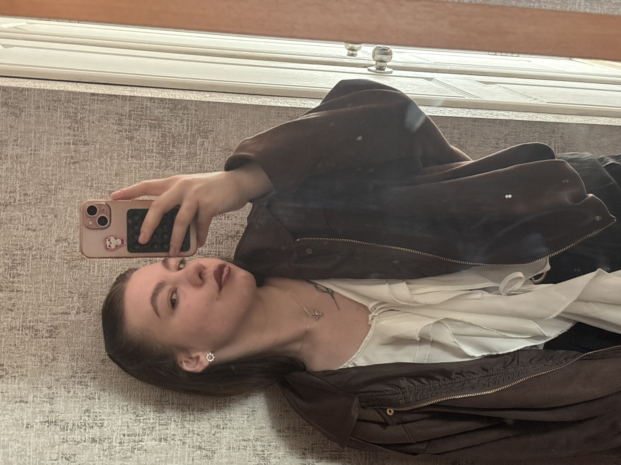 A woman is taking a selfie in front of a mirror. She is wearing a brown jacket over a white top. She is holding a pink iPhone. The background appears to be a wall with a textured pattern and a portion of a door with a silver handle visible above. The location is most likely in Perm, Russia, as implied in the prompt, though there is no direct evidence within the image itself to confirm this.