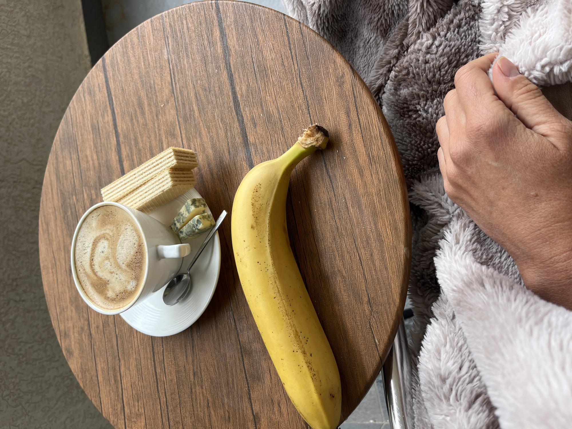 A close-up, top-down shot displays a scene of breakfast or a snack atop a round, wooden-patterned table. A white teacup sits on a saucer filled with coffee, adorned with latte art, a small spoon, wafers, and blue cheese. Next to these treats lies a ripe, yellow banana. A person's hand rests on a grey, fluffy blanket. The setting suggests an indoor environment. The location, based on available information, cannot be confirmed as Budva, Montenegro.