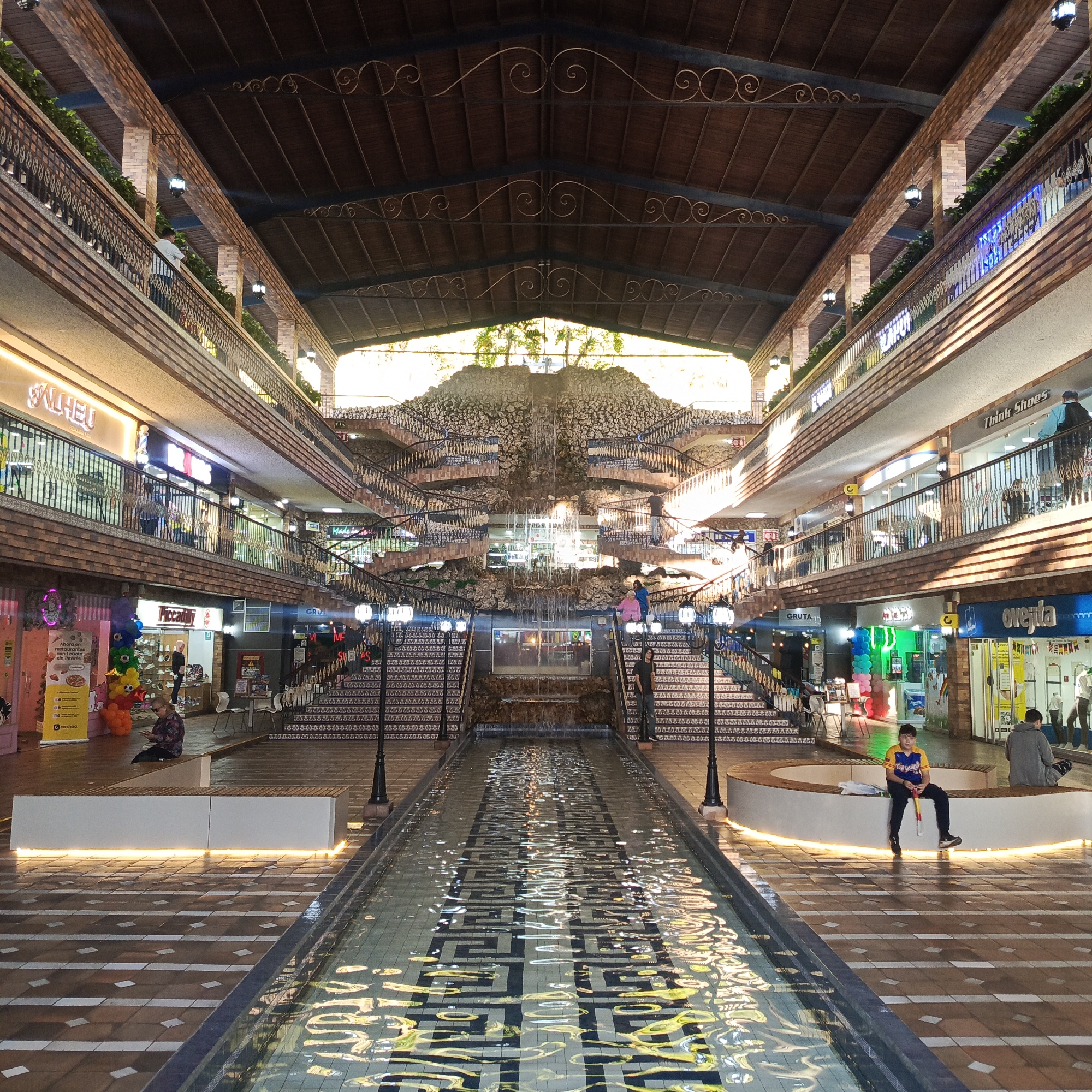 The image depicts the interior of a shopping mall with a unique architectural design featuring a central water feature and multiple levels of shops. The mall appears to be open-air, with a high, vaulted ceiling constructed of wood and metal accents, allowing natural light to filter in from the top. On either side of the central water channel are multi-tiered walkways lined with shops, each with storefronts and balconies overlooking the main area. A large, rock-and-water feature serves as a central focal point, with staircases spiraling up on either side, adorned with potted plants and ornate railings. The flooring is a patterned tile mosaic, and the water channel itself features a geometric mosaic design. Several people are visible throughout the mall, some browsing shops, others sitting on benches, and a few individuals are walking. The overall impression is one of a vibrant, well-lit, and architecturally interesting commercial space. This mall is located in the city of Carrizal, Venezuela.