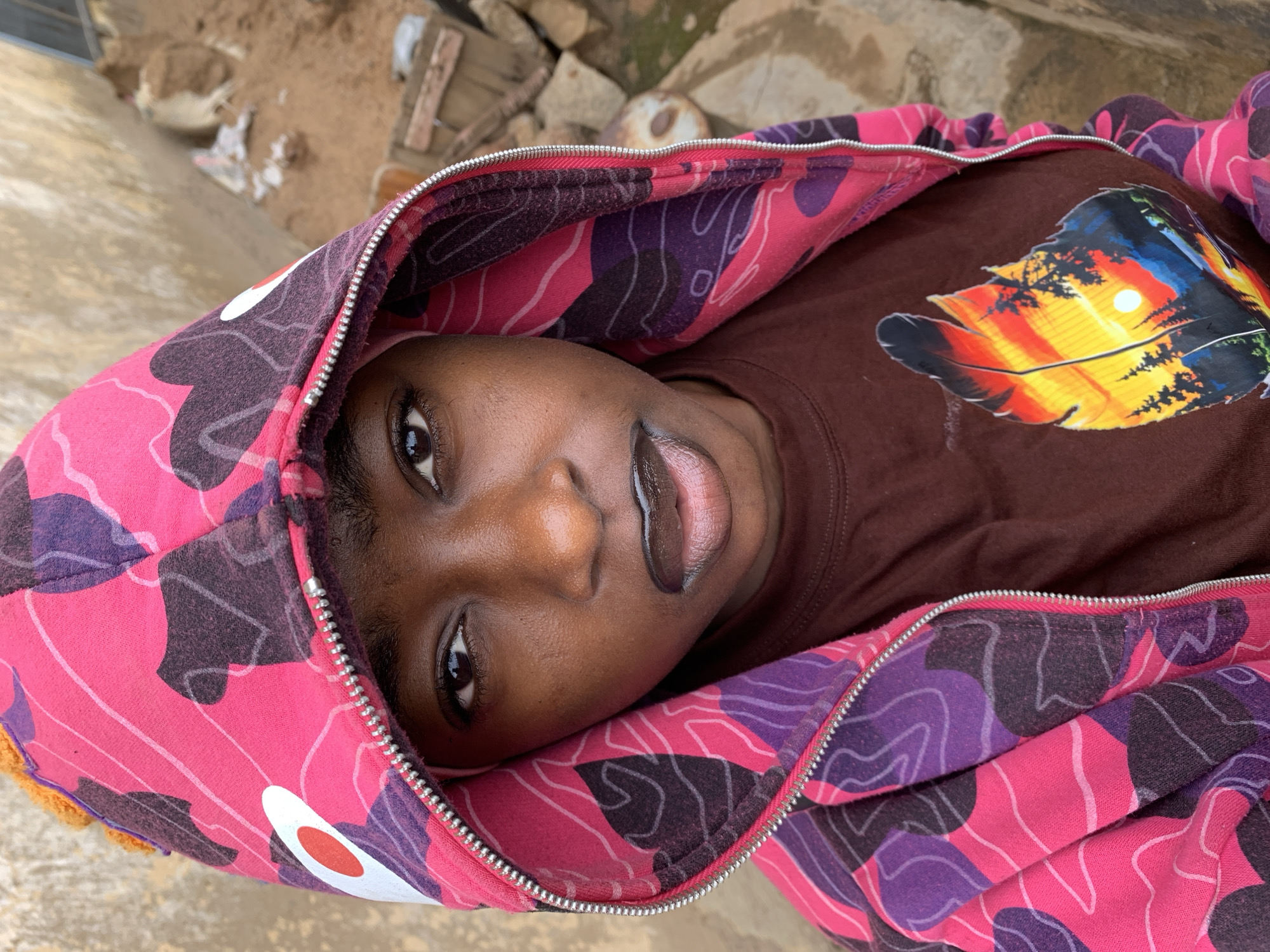A close-up, slightly angled, eye-level photograph depicts a person with dark skin lying down. The person is wearing a brown T-shirt with a brightly colored feather graphic. A pink and purple camouflage-patterned hooded jacket surrounds their face, the hood up. The visible surroundings include a concrete surface and some dirt, debris, and stones. The scene appears to be located outdoors, likely in a residential or semi-urban area within Rafin Par, Nigeria.