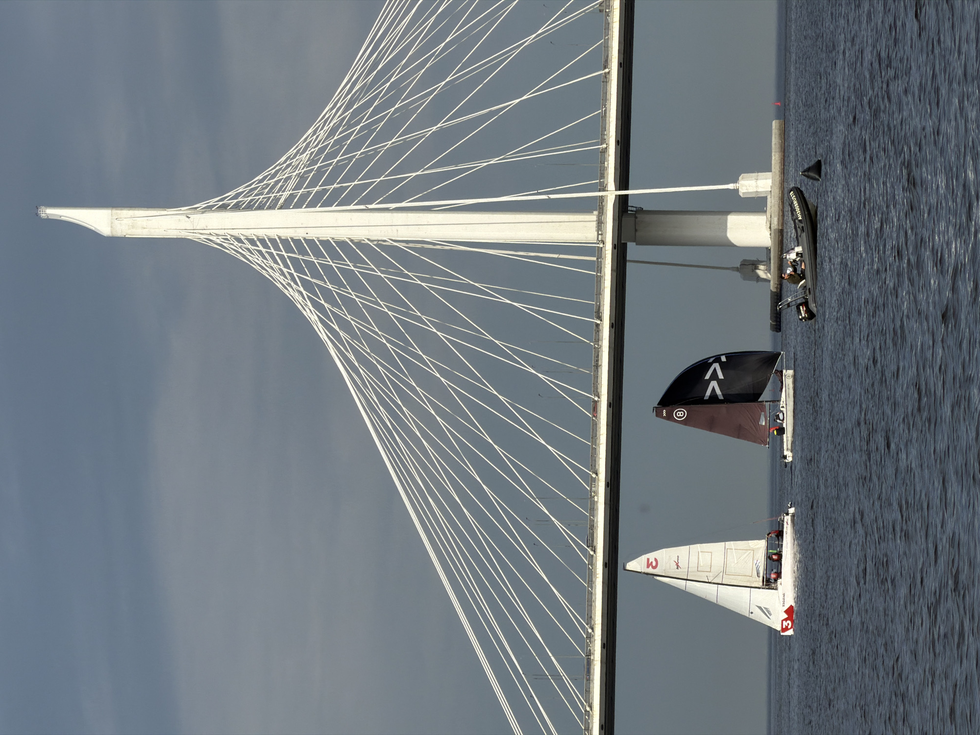 The image captures a scene of sailing boats and a motorboat on the water beneath a large, white cable-stayed bridge. The bridge, a prominent landmark, features a distinctive, tall, white pylon from which numerous white cables extend to support the roadway. The sky is overcast and a muted gray, with a hint of blue.
In the foreground, two sailboats are actively sailing. The sailboat on the left has a white sail with red markings, including the number "3" prominently displayed. The sailboat on the right has a dark, possibly black, sail with white markings, including a symbol that resembles an inverted 'V' or chevron. Both sailboats appear to have people on board, engaged in the activity of sailing.
Further to the right, a dark gray motorboat is moving across the water. A single person is visible at the helm, operating the boat. Next to the motorboat, a dark, triangular object, possibly a buoy or part of a marker, is floating in the water. In the distance, near the bridge supports, a small red marker buoy is visible.
The water is choppy, indicating a moderate wind. The overall impression is one of a regatta or sailing event taking place under a bridge in a coastal or riverine environment. This bridge is likely the Western High-Speed Diameter (WHSD) bridge in Saint Petersburg, Russia, known for its distinctive architecture and its role in connecting different parts of the city across the Neva Bay.