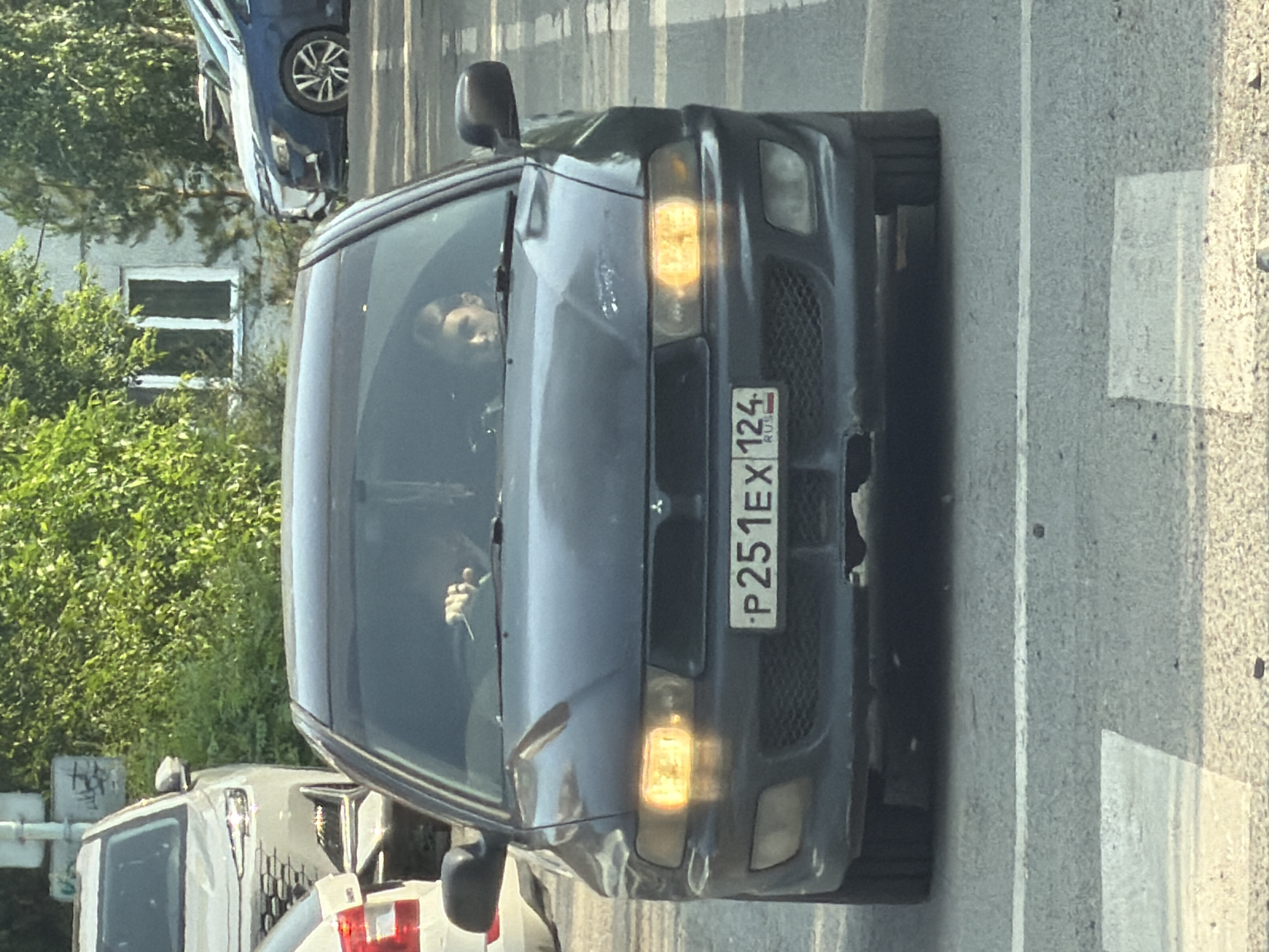 A dark-colored Mitsubishi sedan is shown from the front, driving on a paved road. The car is tilted, with the top of the image pointing downwards. Inside the car, a person is visible behind the steering wheel, with their right arm resting on the door. The person's face is partially obscured by the angle, but they appear to be looking forward.
The car has its headlights on, casting a yellow glow. The license plate is visible and reads "P251EX 124 RUS". The road appears to be an asphalt surface with white lane markings. In the background, to the left, are green trees and a building with windows. To the right, the road surface transitions to a lighter concrete or paved area with white markings, suggesting a pedestrian crossing or a change in road surface.
The lighting suggests it is daytime, possibly late afternoon due to the warm tone of the headlights and the light on the road. The weather appears to be clear. The scene is likely in an urban or suburban environment, given the presence of the road, building, and trees. The overall impression is of a vehicle in motion on a street. There are some visible damages on the front bumper of the car, suggesting a minor collision or impact.