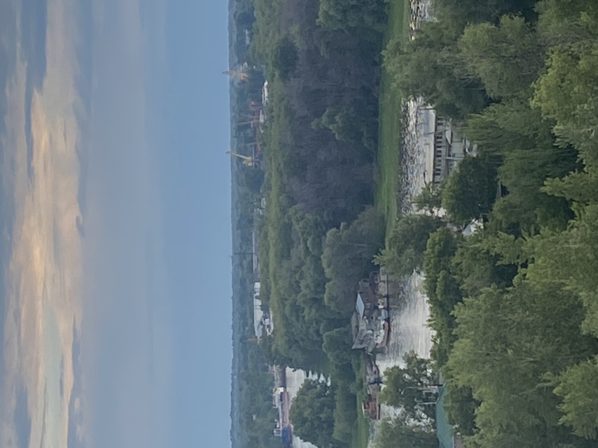 This is an aerial, landscape-oriented photograph of a wide river flowing through a heavily forested area. The river appears to be a busy industrial waterway. Several boats and barges are visible on the water, some moored near the shore and others in transit. On the left bank, there are large industrial structures, including what appear to be cranes or construction equipment, suggesting a port or industrial complex. The dense green foliage of trees lines the banks of the river and extends into the distance, indicating a natural landscape juxtaposed with industrial activity. The sky is partly cloudy, with soft, diffused light suggesting it is either early morning or late afternoon. The overall mood is one of natural beauty coexisting with human industry. No text is visible in the image. The location is identified as Novokuybyshevsk, Russia.
