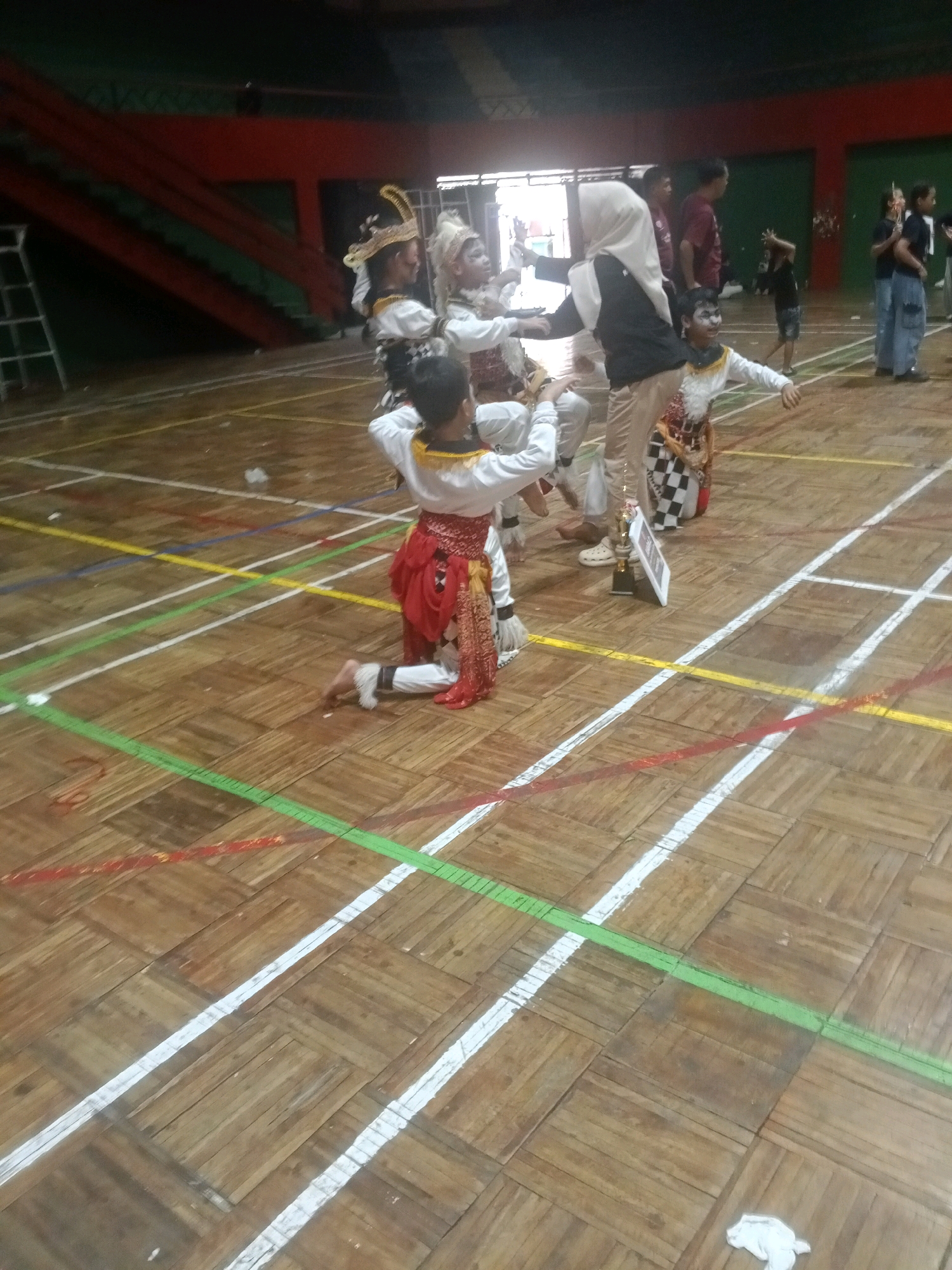 In the gymnasium of Wuryorejo, Indonesia, several young individuals dressed in traditional Indonesian performance attire are engaged in a dance. The participants wear distinctive headpieces, white tops with ornate gold trim, and red skirts or sarongs over white pants. One individual is kneeling on the wooden floor, while others stand. A woman wearing a hijab and a black top is also present, appearing to be an instructor or chaperone. The floor is marked with colored lines, suggesting it is a sports court. In the background, bleachers are visible, and other spectators and participants are scattered around the gymnasium. The lighting is artificial, typical of an indoor sports facility.