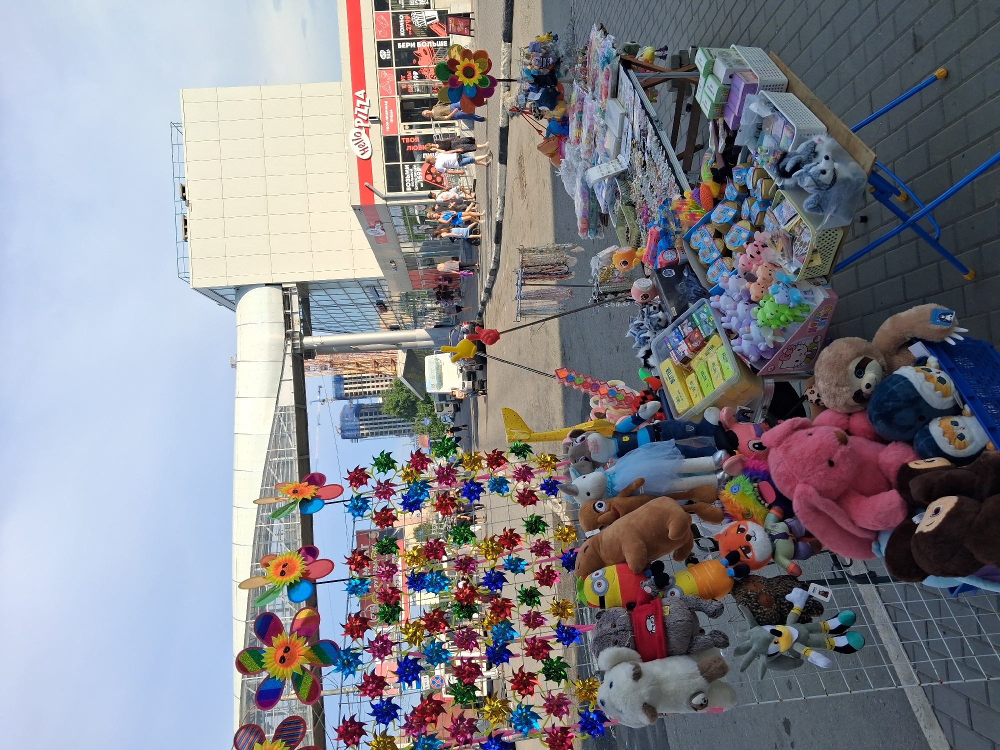 A street market is set up on a paved sidewalk in front of a modern building complex that includes a "Hello Pizza" restaurant. The market displays a colorful array of goods, primarily plush toys and novelty items like pinwheels. Stalls are arranged on tables and metal racks. In the background, a pedestrian overpass crosses the street, and various vehicles, including cars and a bus, are visible. People are walking around the market and on the sidewalk. The sky is blue with some scattered clouds. The scene suggests a typical urban commercial area during daylight hours.