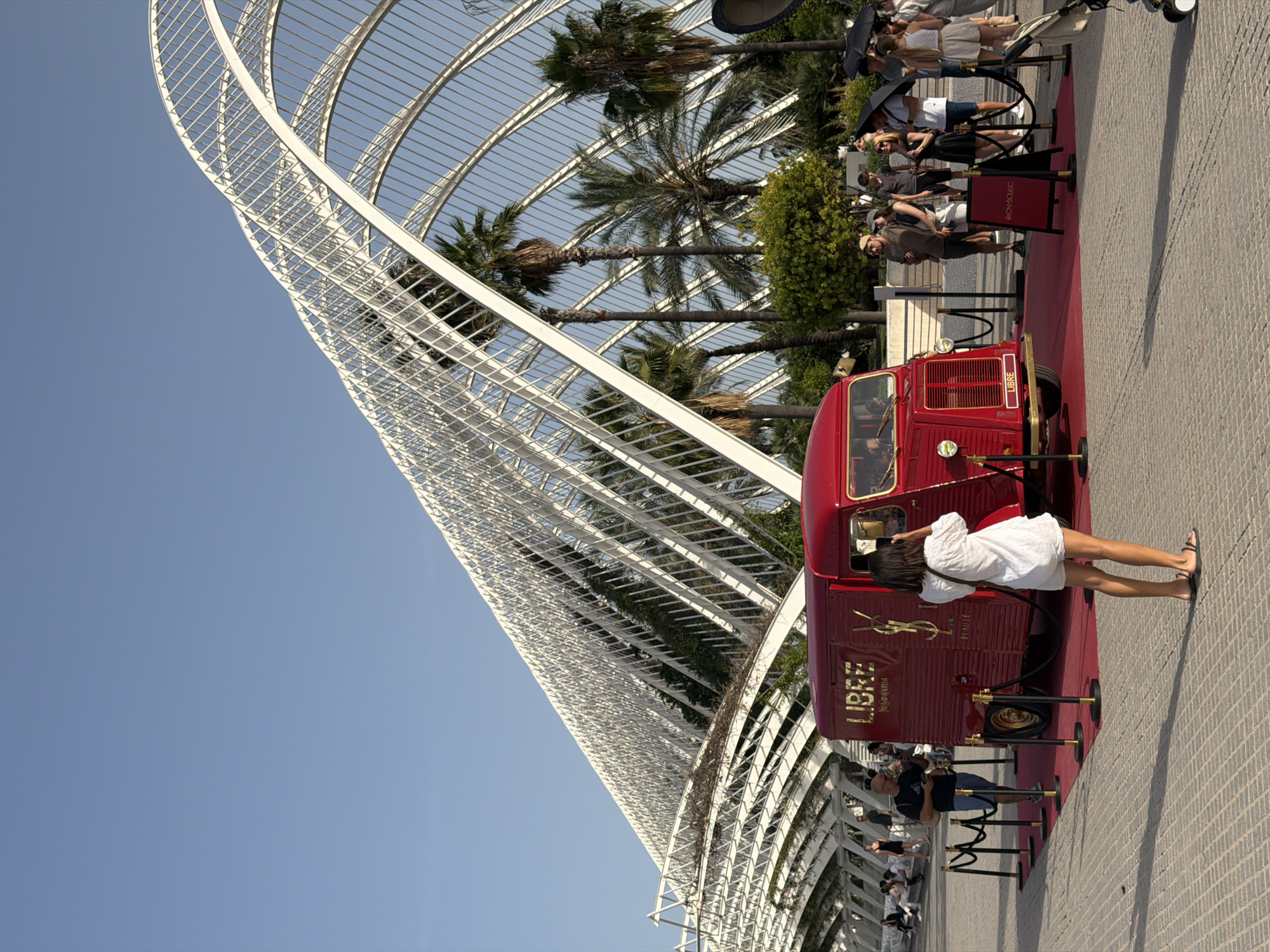 The image depicts a bright, sunny outdoor scene in front of a large, white, arched architectural structure. In the foreground, a classic red van branded with "LIBRE" and the Yves Saint Laurent logo is parked on a red carpet. A woman in a white dress is standing in front of the van, taking a photo with her phone. Several people are gathered around the van, some looking at it and others conversing. A velvet rope barrier separates the van and red carpet area from the onlookers. Palm trees and lush green foliage are visible behind the van, under the arches of the white structure. The ground is paved with textured grey bricks. The sky is clear blue.