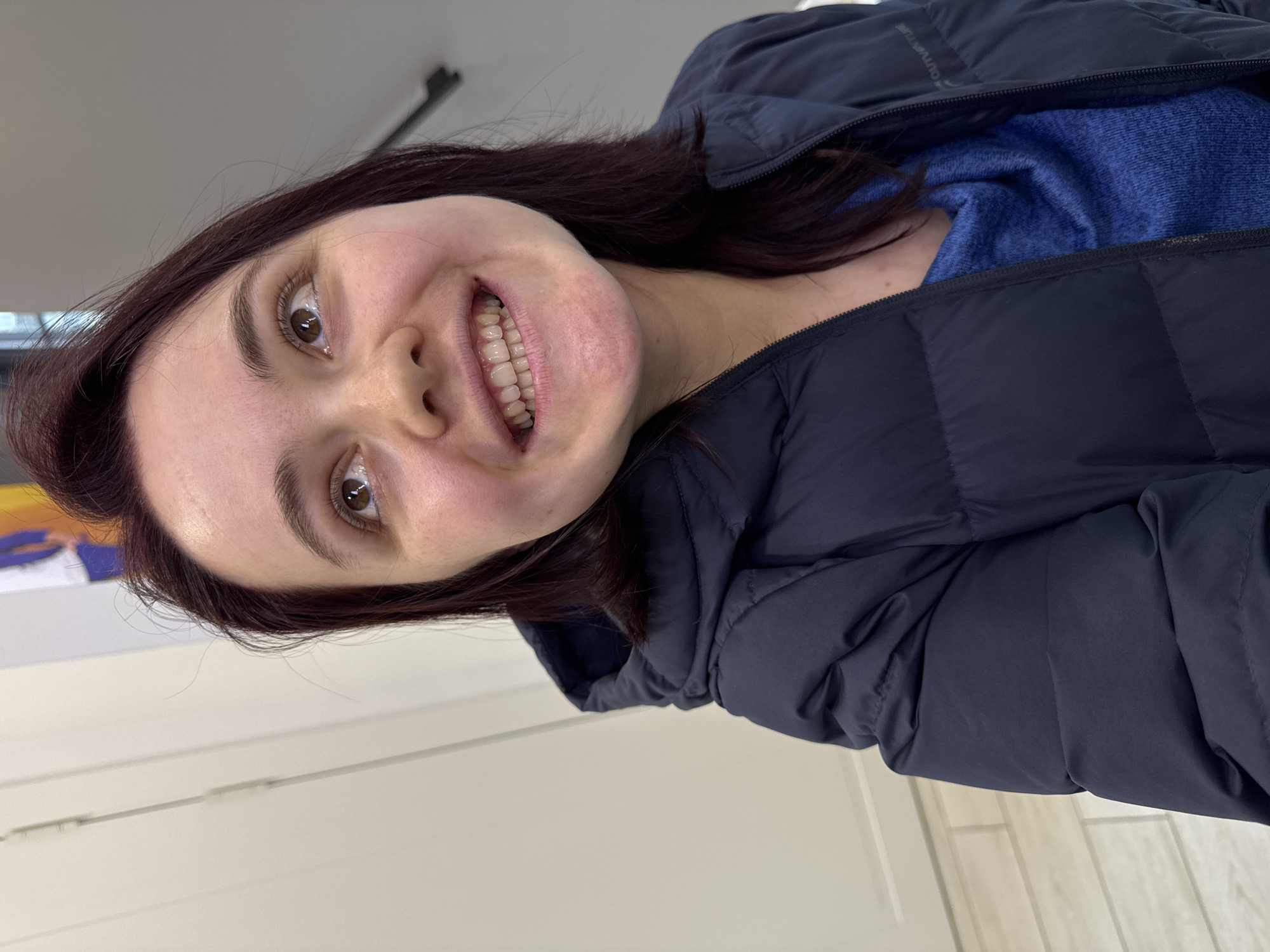 A woman with brown hair is smiling at the camera, wearing a blue sweater and a dark blue puffer jacket. The background is out of focus, with a white wall and a glimpse of a blurred object with yellow and blue colors. The image appears to be taken indoors. There is no specific information to determine the location is in Izhevsk, Russia.