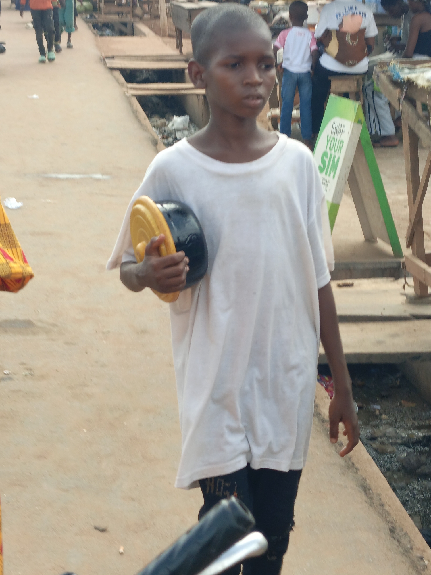 In Akwanga, Nigeria, a young person walks through a marketplace carrying a black and yellow object. The individual wears a white oversized t-shirt and black leggings. The market scene includes people, stalls, and signage. Open drains line the edge of the walkway.