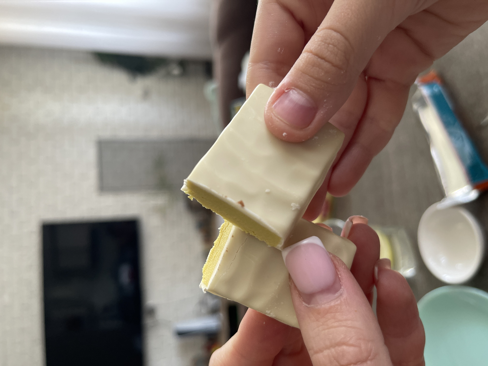 In Blagoveshchensk, Russia, a close-up shot captures two hands holding pieces of a white chocolate confection with a layered yellow filling, likely a cake or cookie. The left hand, with neatly manicured nails, holds one piece, while the right hand grasps another, bringing them together as if to share or break them further. The confection appears to have a smooth, slightly textured white chocolate coating. In the background, out of focus, are elements of an indoor environment, suggesting a home setting. A large black television screen is visible on a wall adorned with white brick-patterned wallpaper. To the right, a white curtain hangs near a window. On the table in the foreground, a light blue dish and a small white bowl are present. The overall scene is casual and domestic, focusing on the dessert being held.
