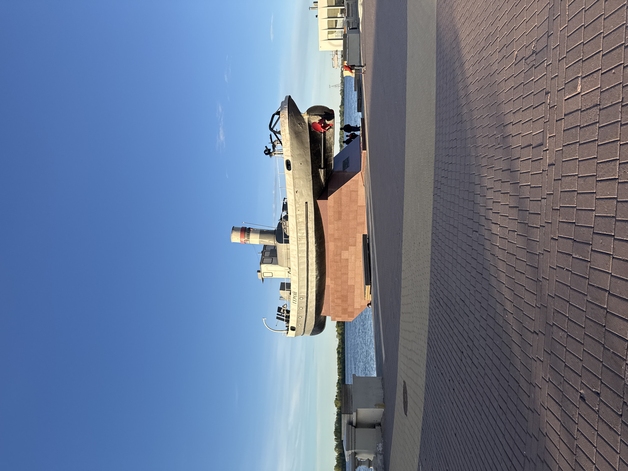 The image depicts the "Hero" gunboat monument, located on the Upper Volga embankment in Nizhny Novgorod, Russia. The gunboat, mounted on a reddish-brown stone pedestal, is positioned parallel to the Volga River. The bow of the boat faces towards the city. The sky is clear and blue. A cobblestone pedestrian walkway runs along the right side of the image. In the background, across the river, are trees and buildings. Several people are visible on the embankment, including one in a bright red shirt standing near the bow of the gunboat.