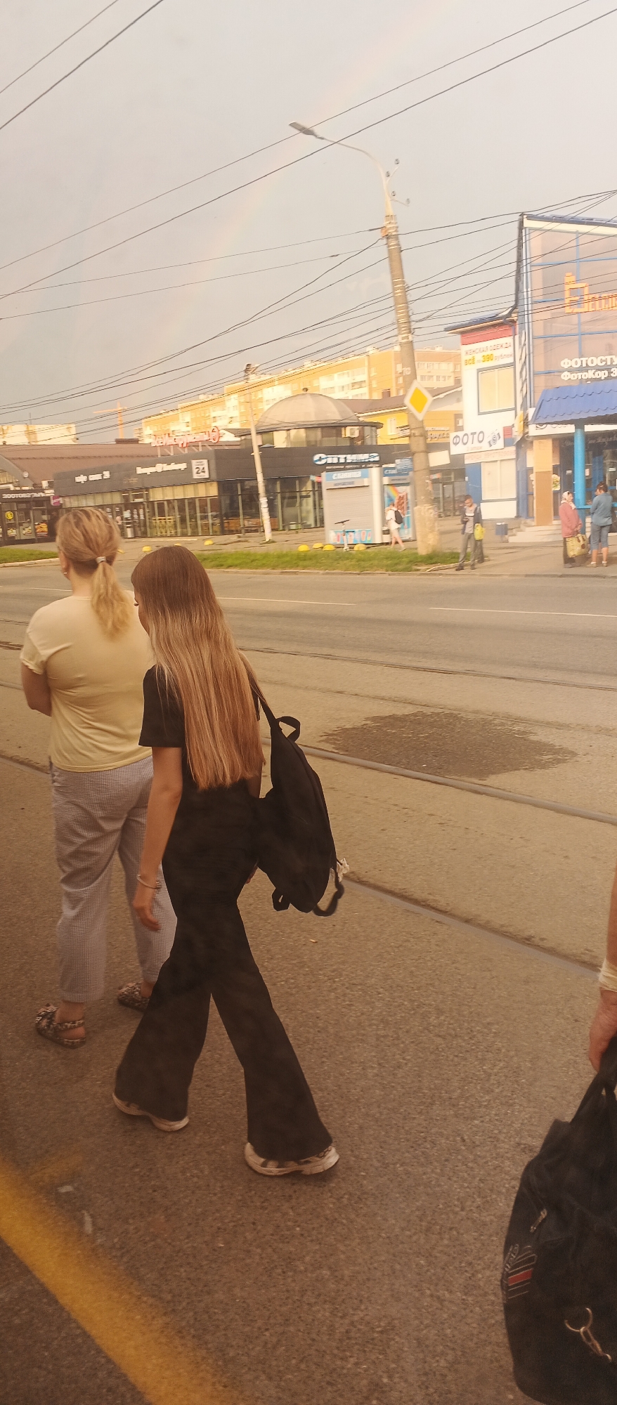 Two women are walking on a sidewalk in Izhevsk, Russia, in front of shops. The woman in the foreground, on the right, has long blonde hair and is wearing a black t-shirt and black bell-bottom pants. She is carrying a black backpack. The woman behind her, on the left, has blonde hair in a ponytail and is wearing a light-colored t-shirt and gray striped pants. She is wearing sandals. In the background, there are several buildings with storefronts, including a "Фотостудия" (Photo Studio) and shops selling clothing. A rainbow is visible in the sky. The scene appears to be a street in a commercial area of the city.