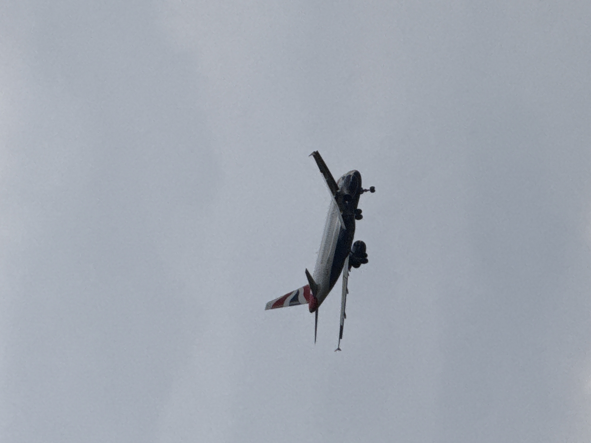 An image captures a large passenger airplane performing a dramatic, near-vertical banking maneuver against a uniformly overcast, light grey sky. The aircraft, identified as a British Airways plane by the distinct red, white, and blue Union Jack livery visible on its tail fin, is seen from below. Its nose is angled sharply upwards and slightly to the left, while the tail points downwards and to the right, indicating an extreme angle of ascent or turn.
The plane's underside and left side are partially visible, revealing several engine nacelles and what appear to be deployed landing gear struts and wheels. The cloudy, featureless sky suggests an overcast day. The extreme orientation of the aircraft implies it is engaged in an airshow performance or a highly unusual maneuver, rather than standard flight. The setting for this event is Dolni Bogrov, Bulgaria. No explicit text is visible beyond the recognizable tail livery.