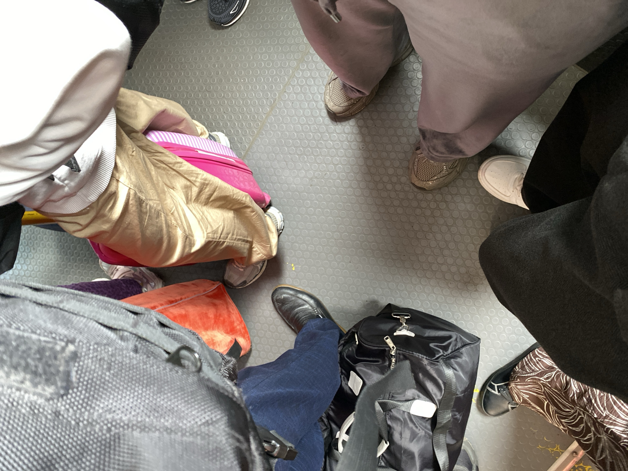 The image shows a bird's-eye view of people's legs and luggage on a grey, textured floor, likely inside a mode of transportation. Several individuals are visible, distinguished by their clothing and footwear. One person wears dark blue trousers and black leather loafers, standing next to a large black duffel bag. Another person is dressed in gold-colored pants and a white sweatshirt, with a bright pink suitcase beside them. A third individual wears dusty rose-colored trousers and light brown sneakers. Further to the left, a person is seen with patterned brown and white trousers and black shoes with white accents, adjacent to a large black backpack. The floor has a repetitive circular pattern, characteristic of some bus or train interiors. There is no specific information in the image to confirm the location as Новое Мартьяново, Russia.