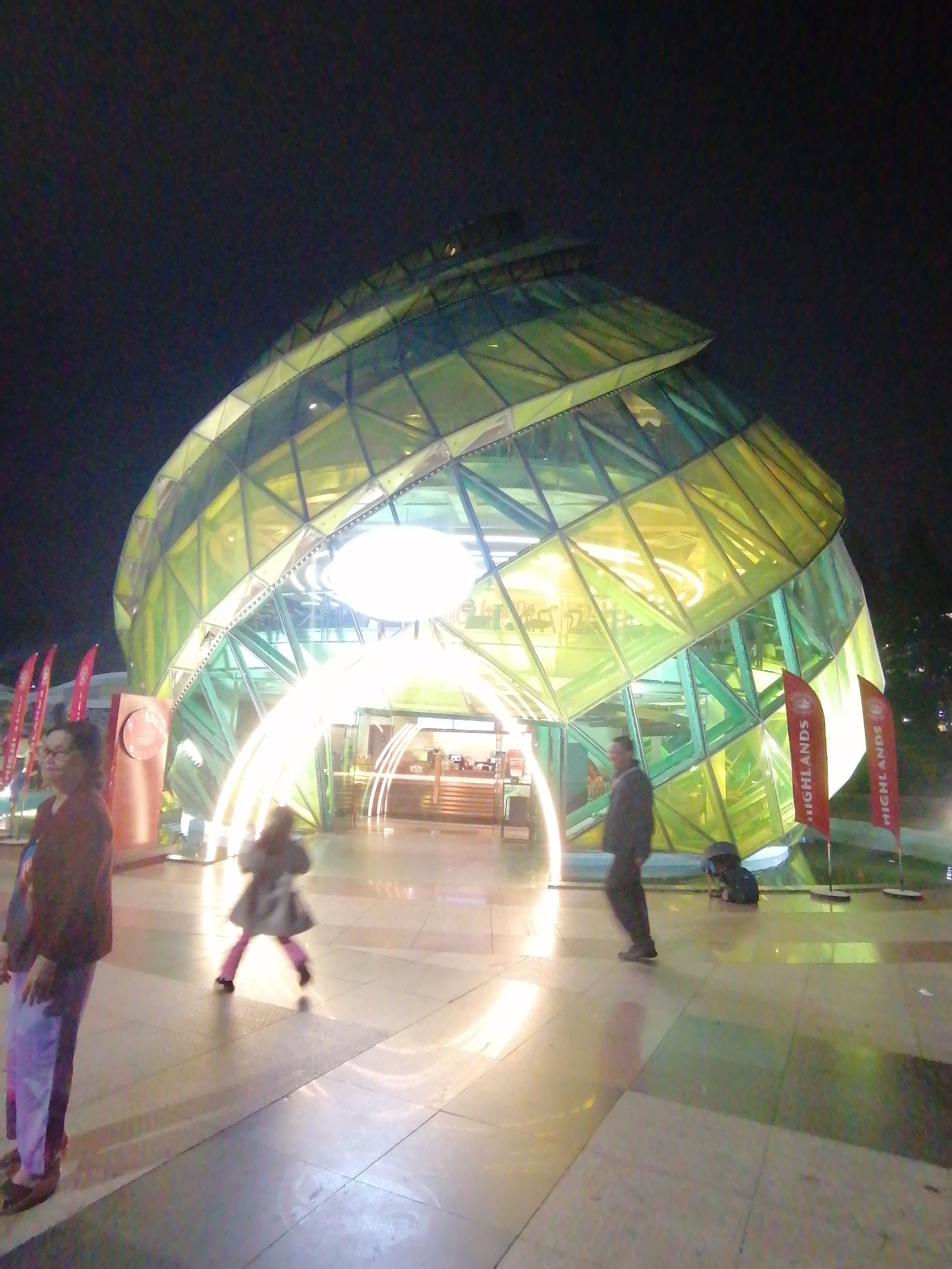 At night, a building with a dome-shaped structure featuring triangular window panes stands illuminated. A circular light fixture glows intensely in the entrance. A woman with glasses and a maroon top stands to the left. A child wearing a coat walks through the entrance. A man in a suit walks to the right of the entrance. A backpack sits on the ground on the right. Red flags are placed in front of the building, bearing the name "HIGHLANDS". The location is the Da Lat city, Vietnam.