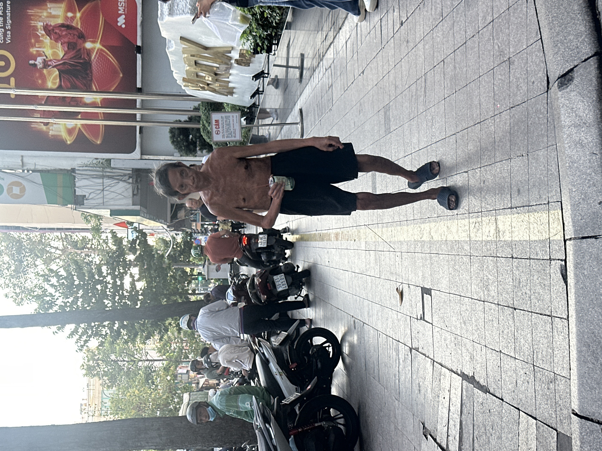 An outdoor street scene in Thủ Đức, Vietnam, is depicted during daytime. In the central foreground, a shirtless, lean man with dark hair and black shorts walks towards the viewer's left, holding a green and white can in his right hand and wearing dark grey sandals. To his right, a partially visible individual wears blue jeans and white sneakers, holding a smartphone and a clear plastic bag containing a green liquid. On the left side of the frame, multiple individuals are present on or near motorcycles parked on the sidewalk. One person, wearing a green jacket, helmet, and blue face mask, sits on a white scooter. Another individual, dressed in a white long-sleeved shirt and helmet, stands near a motorcycle displaying the license plate "59-E1 485.09." In the background, a building features a large vertical advertisement showcasing a woman in a red gown with "MSB Visa Signature" branding. Below this advertisement, a white sign with red Vietnamese text "CÁM" (meaning forbidden) is visible, adjacent to a large structure with reflective gold lettering. The ground consists of a tiled sidewalk with a yellow stripe, bordered by a concrete curb. The scene is illuminated by bright sunlight, casting distinct shadows.