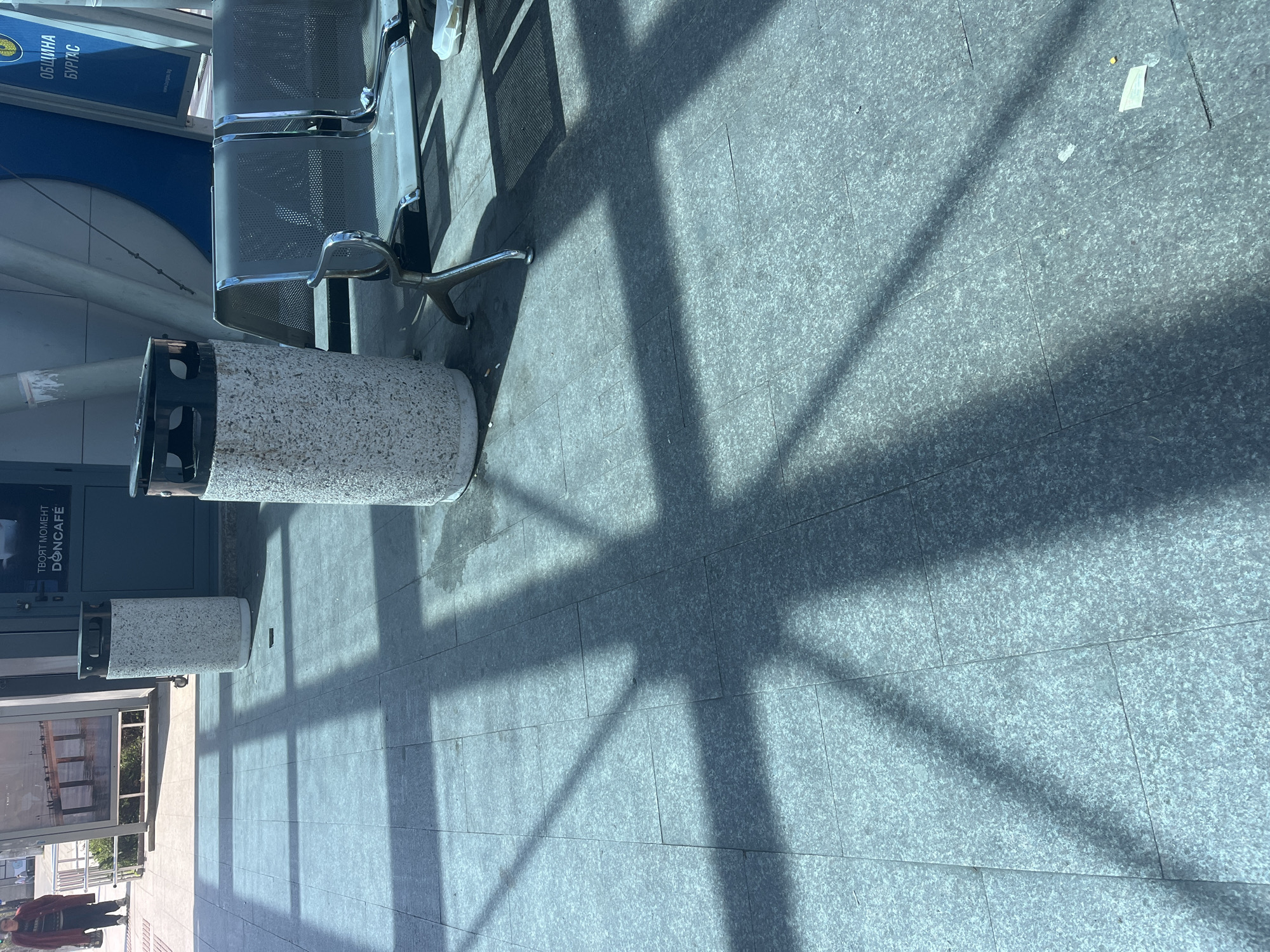 The image depicts a public waiting area, possibly part of a transportation hub or a large commercial building, in Burgas, Bulgaria. The setting is characterized by a light grey, tiled floor with a speckled appearance, likely granite or similar stone. Strong, diagonal shadows from a large window or roof structure dominate the right half of the frame, indicating bright sunlight and a clear day.
In the mid-ground, a two-seater metal bench with perforated seats and backs, typical of public waiting areas, is visible. To the left of the bench, two round, concrete-textured trash cans with black tops are placed. The closest trash can is in the foreground, showing some minor wear.
On the far left, a vertical blue sign or banner is present, featuring white Cyrillic text that reads "ОБЩИНА БУРГАС" (Municipality Burgas) and a web address "www.burgas.bg", confirming the location. Below this sign, partially obscured, is another sign with the text "ТВОЯТ МОМЕНТ DONCAFE" (Your Moment DONCAFE), suggesting a cafe nearby.
A person, largely obscured and wearing dark clothing, is partially visible in the bottom left corner, walking or standing in the bright sunlight. The lighting suggests it is daytime, possibly late morning or early afternoon, given the sharp, defined shadows. The overall scene conveys a functional and utilitarian public space.