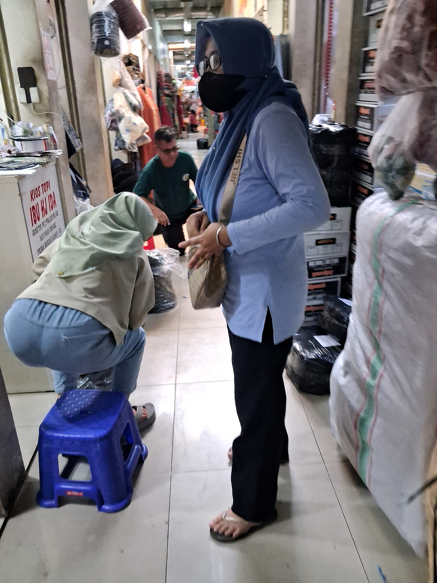 The image captures a scene inside a market stall in Surakarta, Indonesia. Two individuals are partially visible. On the left, a person wearing a light green hijab and light blue pants is crouched down, their back to the camera. They are seated on a small, blue plastic stool. To the right, a person wearing a blue hijab, glasses, and a black face mask is standing. They have on a light blue long-sleeved shirt and black pants, and are carrying a beige handbag. A man wearing a green shirt and sunglasses is visible in the background. The environment appears to be a retail space with shelves of goods and clothing items hanging in the background. The flooring is tiled.