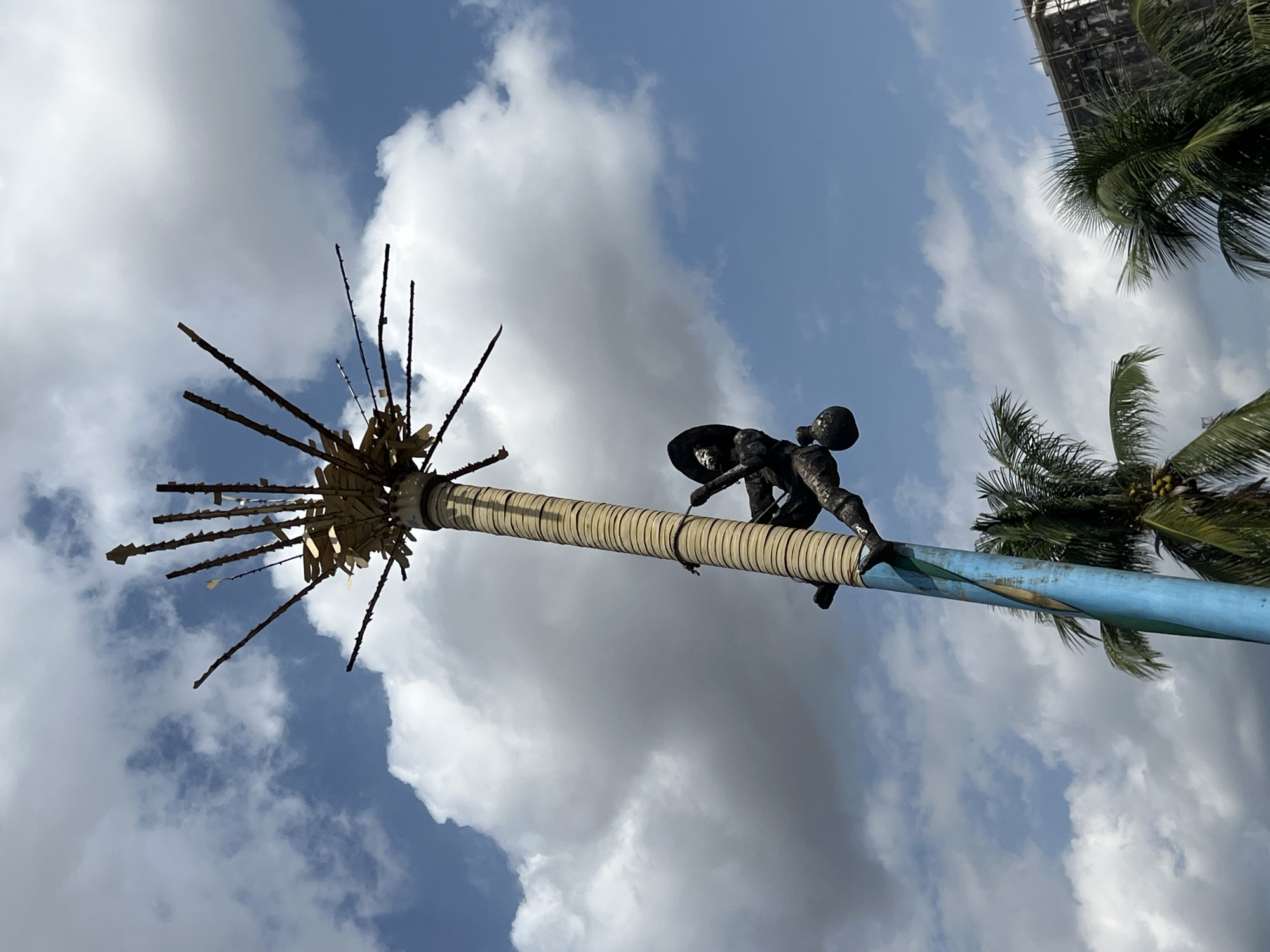 The image, captured from a low angle, presents a tall, decorated pole angled diagonally upwards against a bright blue sky with scattered white clouds.
The pole is composed of two distinct sections. The lower section, positioned on the right side of the frame, is a weathered blue with faded painted motifs that appear to depict a person and palm trees. The upper section, to the left, is wrapped tightly with numerous light-colored horizontal bands, resembling rope or woven material. Crowning the far left end of this section is a spiky, radiating structure with dark projections and smaller white elements attached, possibly serving as a decorative or symbolic finial.
A dark-colored, sculptural figure is attached to the blue section of the pole. The human-like figure, appearing to be climbing or clinging to the pole, is rendered in a dynamic pose and wears a distinct wide-brimmed hat or head covering.
In the background, several green palm trees are visible on the right side, suggesting a tropical setting. Behind these trees, a building under construction, identifiable by its visible scaffolding, is partially glimpsed at the top right.
The scene is brightly lit by daylight, indicating a clear and sunny day with patchy cloud cover. There is no visible text in the image. The structure likely holds cultural or artistic significance within its Onitsha, Nigeria, context.