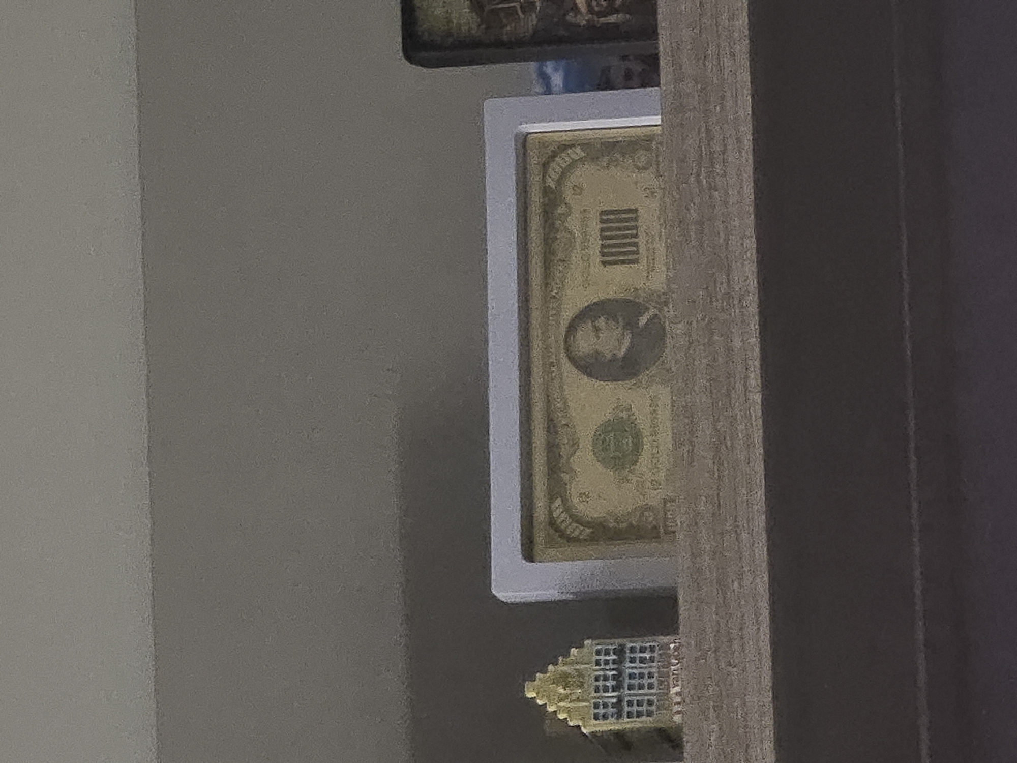 The image displays a close-up, rotated view of decorative items placed against a wall and next to a wooden surface.
**Subjects:**
The central subject is a rectangular, golden-colored bill or commemorative note, enclosed in a white frame. Below this, partially visible, is a small, golden, architectural figurine, possibly representing a building or monument. To the right of these items is a vertical wooden panel or piece of furniture. The background consists of a light gray wall.
**Activity or event taking place:**
No specific activity is occurring; the image captures a static display of objects.
**Notable details:**
The framed golden note is prominently detailed. It features the number "1000" vertically oriented near the top right (when viewed in portrait). A portrait of a person is visible in an oval frame on the left side, and a circular emblem with "100" inside it is seen towards the bottom center. The note appears to be decorative rather than functional currency. The small golden figurine below it has intricate detailing, including what look like multiple small windows or sections. The wooden panel next to the display has a visible grain. The lighting suggests an indoor setting.
**Visible text:**
The most clear text is "1000" on the golden note. A smaller "100" is also visible within a circular emblem on the note. Faint, possibly decorative text is present on the golden figurine, with parts of "YORK" or a similar word discernible on its side.