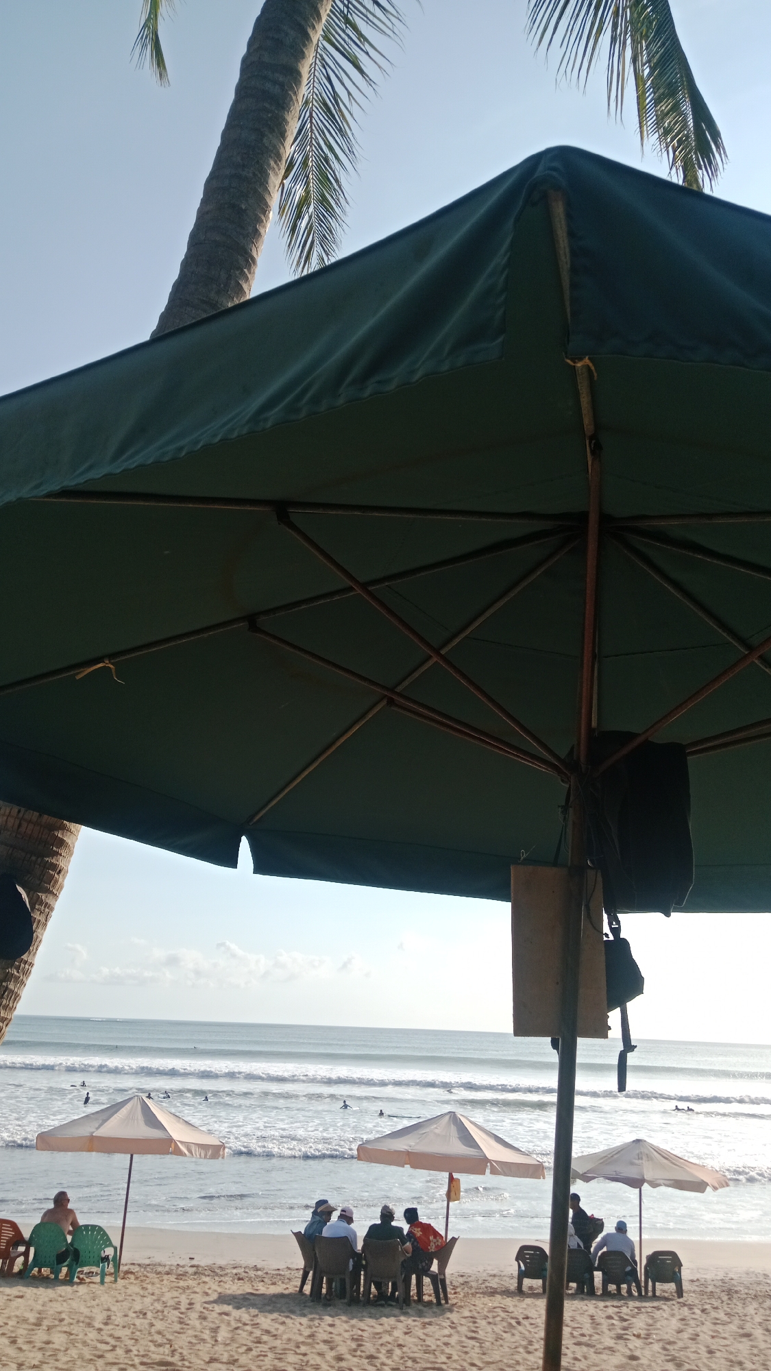 A wide-angle shot captures a beach scene in Kuta, Indonesia, on a bright, sunny day. In the foreground, the underside of a large, dark green beach umbrella dominates the top half of the frame, its skeletal structure of wooden poles visible. To the left, a portion of a palm tree trunk and fronds can be seen against a clear, pale blue sky.
The middle ground of the image reveals a sandy beach with several small groups of people seated at tables under lighter-colored beach umbrellas. On the left, two individuals are seated on plastic chairs facing the ocean, with one person wearing a green shirt. A cluster of five people are seated around a table under a beige umbrella, some appearing to be engaged in conversation. Further to the right, another group of three people are seated under a similar beige umbrella. Several empty plastic chairs are also scattered on the sand.
The background features the ocean with gentle waves breaking and rolling towards the shore. The water is a light blue-green color. Several surfers can be seen dotted across the ocean, some riding waves, while others are paddling. The horizon line is clearly defined, with a hint of clouds in the distance. The overall atmosphere suggests a relaxed and leisurely day at the beach.