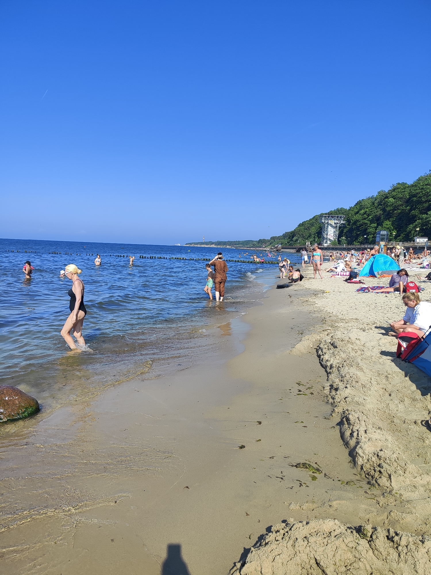 The image depicts a crowded beach on a sunny day in Pionersky, Russia. The sky is clear and blue. The foreground shows a sandy beach with gentle waves lapping at the shore. A large rock is visible in the lower left corner, partially submerged in the water. Several people are in the water, swimming and wading. On the beach, many people are sunbathing, sitting on towels, and relaxing under a blue tent. Some individuals are walking along the shoreline, and children can be seen playing. In the background, a lush green hillside rises, with trees and some structures, including what appears to be a tower-like building. The overall scene conveys a lively atmosphere of a summer day at the seaside.