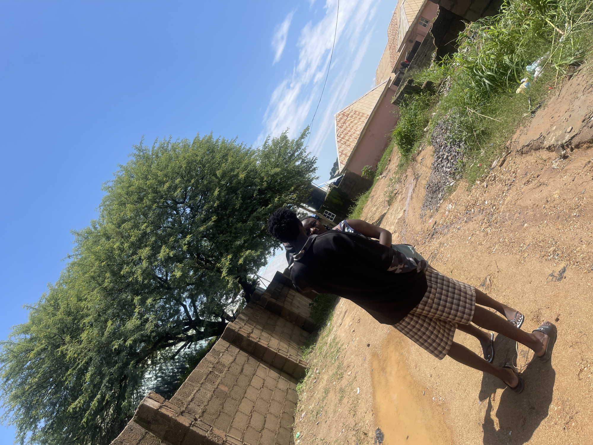 In Rafin Par, Nigeria, a couple stands close together on a dirt path. The male, wearing a black shirt and plaid shorts, embraces the female, who is dressed in a dark top. Behind them, a large tree with green foliage towers against a clear blue sky dotted with white clouds. A brick wall runs along the left side of the frame, and further in the distance, buildings with corrugated roofs are visible. The ground is uneven, with patches of dirt, grass, and a puddle of muddy water.
