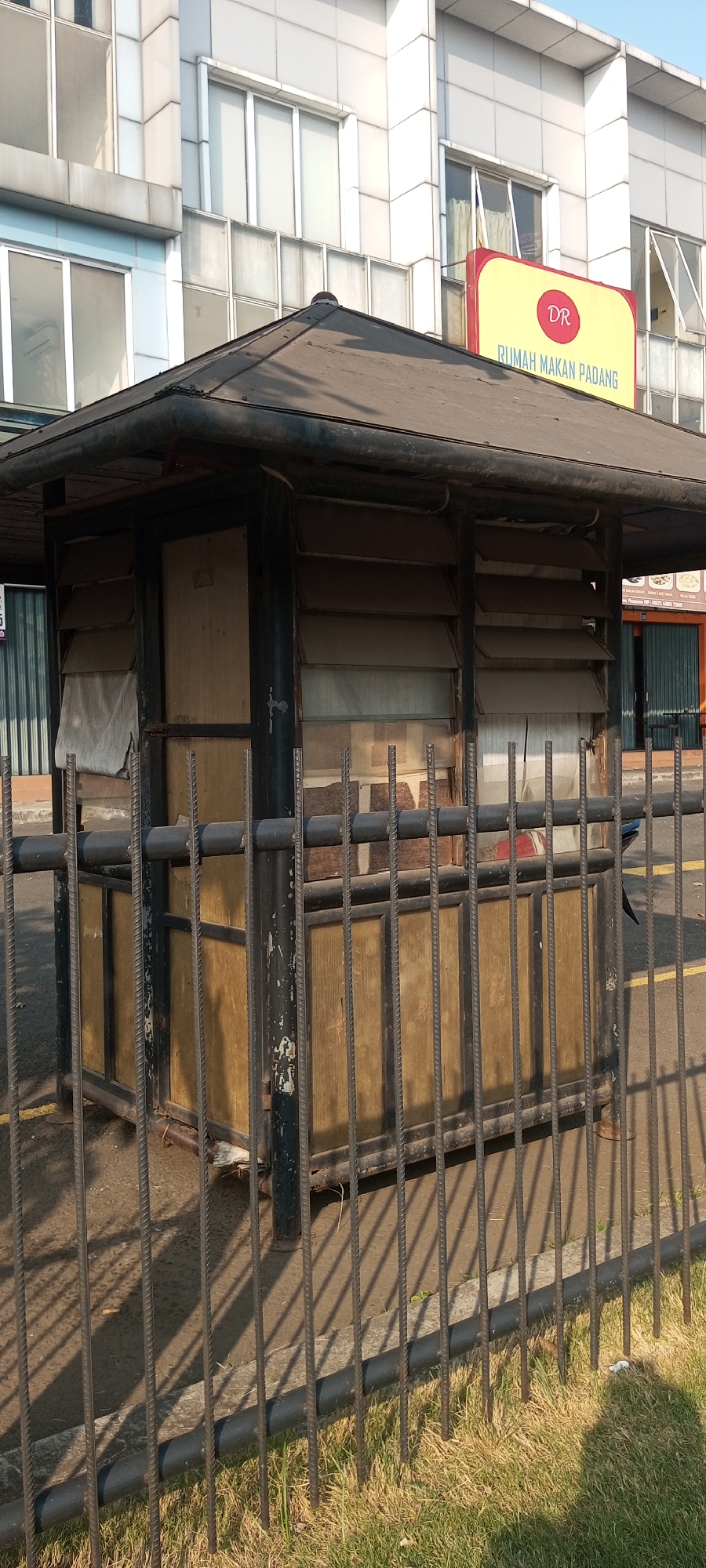 The image depicts a small, weathered guardhouse or kiosk situated behind a metal fence in an urban environment. The structure has a dark, angled roof and dark-colored vertical supports. The walls of the kiosk appear to be made of glass panels with horizontal slats, some of which are covered by a light-colored fabric. There is a wooden door on the front of the kiosk. The metal fence in front of the kiosk is made of vertical rebar-like bars. Behind the kiosk, a modern building with glass windows is visible. A yellow sign with red lettering that reads "DR RUMAH MAKAN PADANG" is attached to the building. The ground in front of the fence is covered with dry grass and a paved area. The scene is lit by sunlight, casting shadows from the fence and kiosk onto the ground. The location is identified as Lambangjaya, Indonesia.
